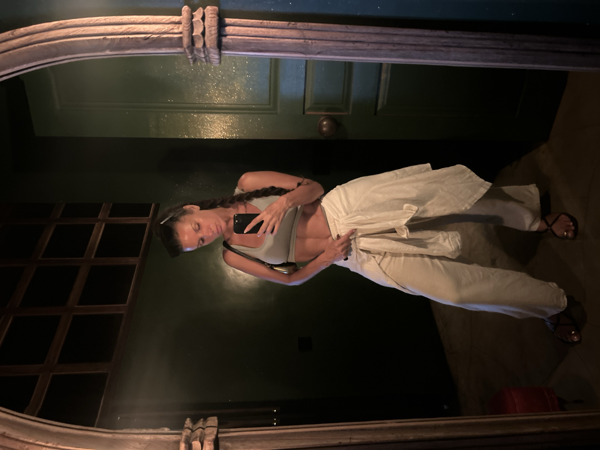 A woman is taking a mirror selfie in what appears to be a dimly lit room with dark green walls and a large, ornate mirror frame. She is wearing a light grey crop top, loose cream-colored pants tied at the waist with a white sash, and black sandals. Her hair is styled in two braids, and she is holding a smartphone in her right hand to capture the image. The background includes a dark, grid-patterned window on the left and a green door with a brass knob on the right. The floor is tiled, and a red stool is partially visible in the reflection on the left.