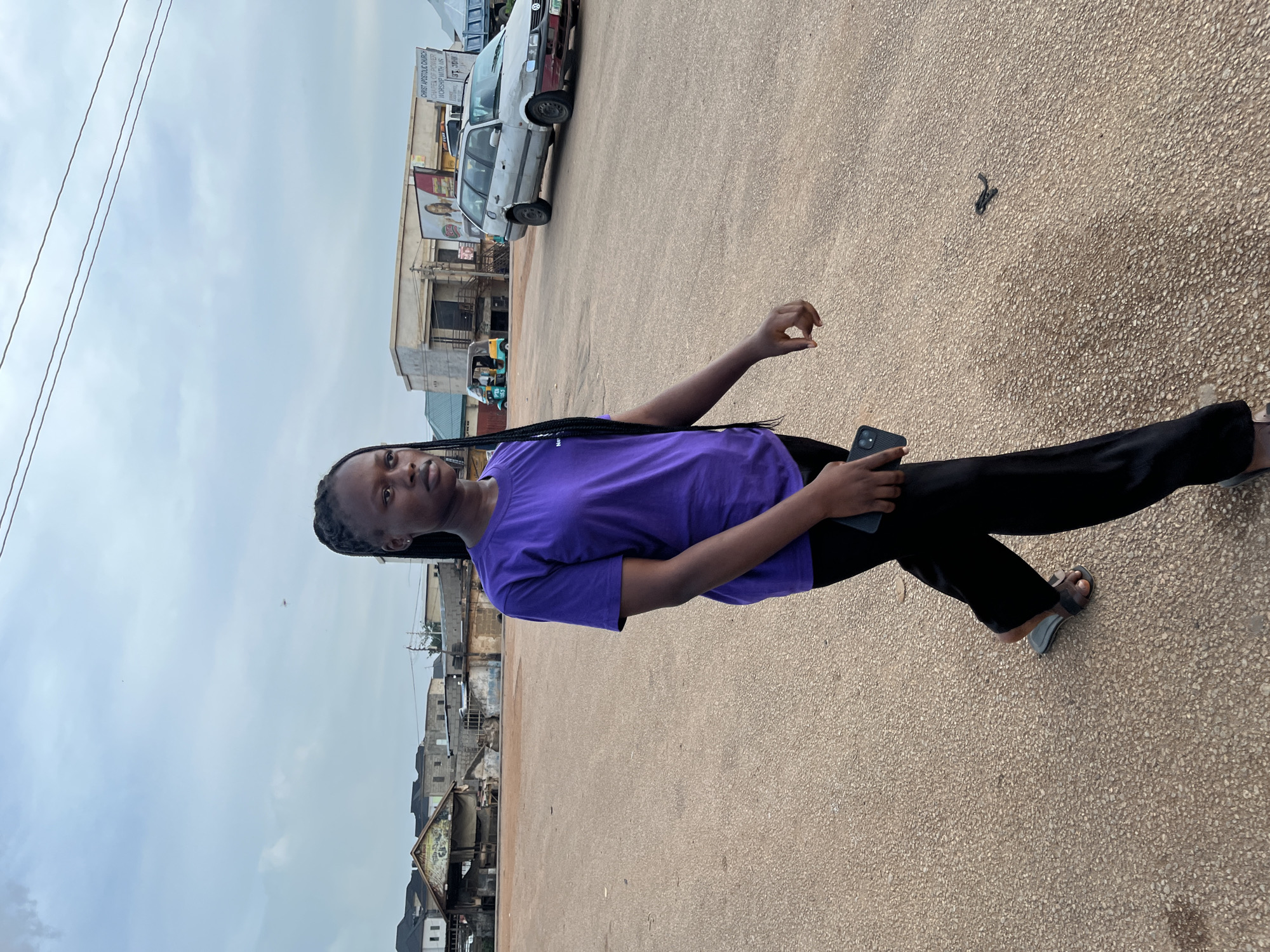 The image, taken in Rafin Par, Nigeria, is rotated 90 degrees clockwise, depicting a young individual standing upright on a paved street or open area. The subject, a person with dark skin and braided hair, wears a bright purple t-shirt and black trousers, along with light-colored sandals. They are holding a dark-colored smartphone in their left hand and making a slight gesture with their right hand, looking directly towards the viewer with a neutral expression.
The ground is a textured, light brown or beige asphalt/concrete mixed with small aggregate stones. In the background, an urban or semi-urban scene unfolds under an overcast sky. Several buildings, appearing to be a mix of residential and commercial structures, line the street. Parked cars are visible, including an older model white sedan. Overhead, power lines traverse the sky, supported by utility poles.
A prominent sign in the background reads: "CHRIST APOSTOLIC CHURCH," with additional text partially visible including "CHURCH OF POWER" and "WORSHIP WITH US." Other fragmented words like "JST." and "TO" can also be discerned on the sign. The overall atmosphere suggests a typical daytime street scene, with muted lighting due to the cloudy weather. A small, dark, twisted object (possibly a piece of debris or a cable tie) lies on the ground near the subject's feet.