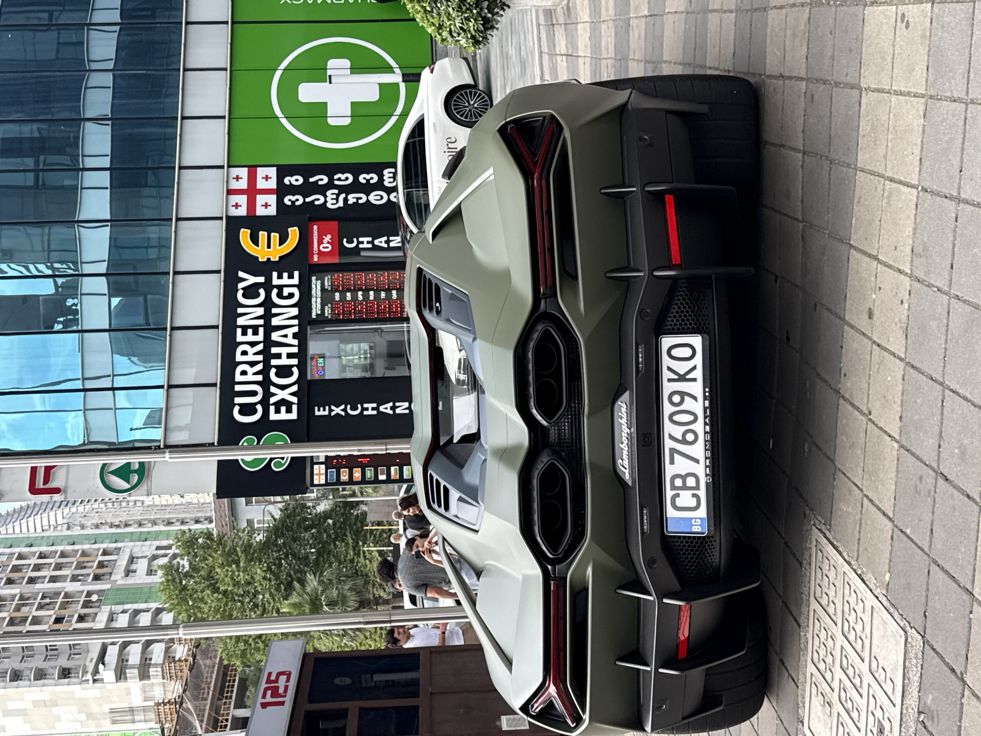 An olive green Lamborghini Aventador SVJ is parked on a tiled city street in front of a building with a "Currency Exchange" sign. The car has a prominent rear spoiler and quad exhaust pipes. The license plate reads "CB 7609 KO" and has a Bulgarian flag emblem. In the background, people are walking, and a white Mercedes-Benz sedan is parked behind the Lamborghini. The building features a green facade with a white cross logo and the word "PHARMACY" visible. The scene appears to be in an urban environment with high-rise buildings under construction visible in the distance.