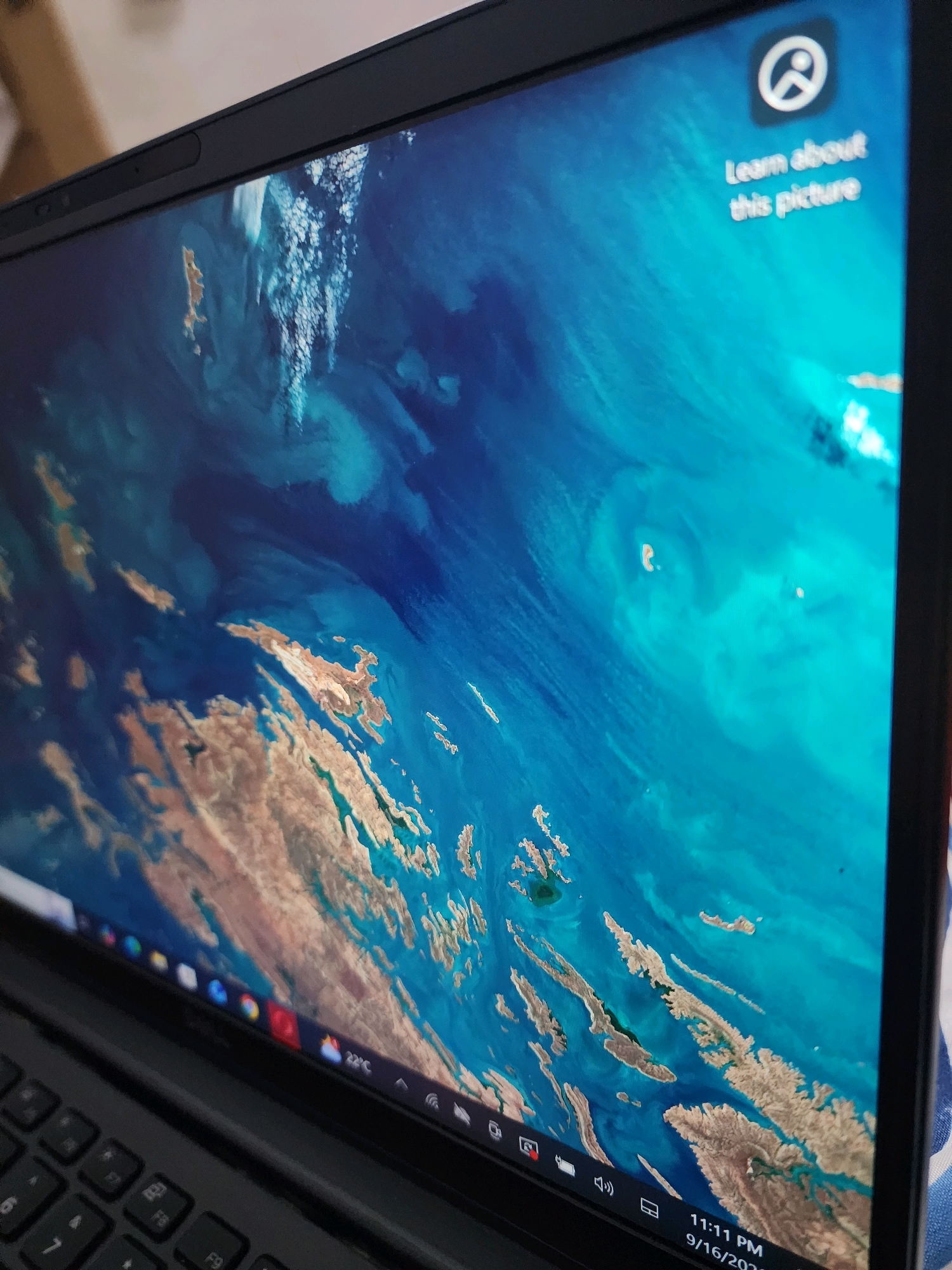 The image shows a close-up of a laptop screen displaying a satellite image of an archipelago. The wallpaper features a vibrant blue ocean dotted with islands that appear to be tan and brown. There is a small pop-up window on the top right of the screen with an icon and the text "Learn about this picture." The laptop's keyboard is visible at the bottom, along with the taskbar which shows the time as "11:11 PM" and the date as "9/16/2020." The laptop is positioned on a surface covered with a blue and white striped fabric. The overall scene suggests a digital interface being viewed in a domestic or personal setting. There is no information in the image that can be used to determine its location in Chamba, Pakistan.