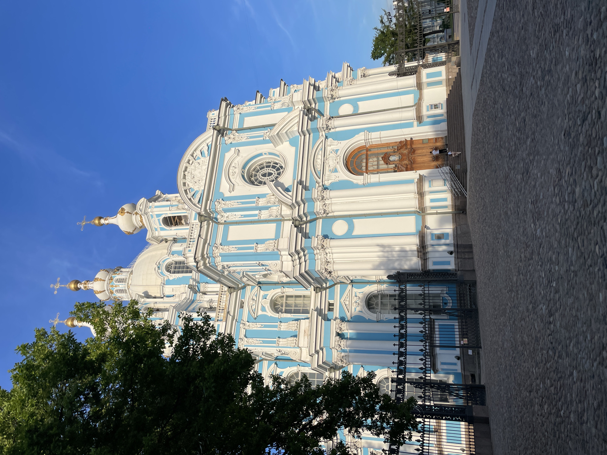 This image depicts the Smolny Cathedral in Saint Petersburg, Russia, under clear blue skies. The prominent structure is a large Baroque-style church, characterized by its striking white and light blue facade, adorned with numerous columns, pilasters, and intricate stucco details. Five golden onion-shaped domes, each topped with an Orthodox cross, rise above the white roofline. Arched and circular windows are distributed across the building's exterior.
In the foreground, a dark gray cobblestone plaza occupies the lower portion of the frame, with some areas appearing to be in shadow. An ornate, dark metal fence with decorative finials runs horizontally across the mid-ground, acting as a boundary for the church grounds. One section of the fence is open, forming a gate.
On the right side of the main entrance, a wide set of stone stairs leads up to a large, arched wooden door. Two individuals are visible on these stairs: one person in a white top and dark pants stands at the top landing, facing the door, while another person in light-colored clothing ascends the lower steps.
A large, leafy green tree partially obscures the left side of the cathedral, casting shadows onto the ground and the lower portion of the building. The sky above is a vibrant, cloudless blue, indicating a bright, sunny day. Shadows cast by the architecture suggest either early morning or late afternoon sunlight.