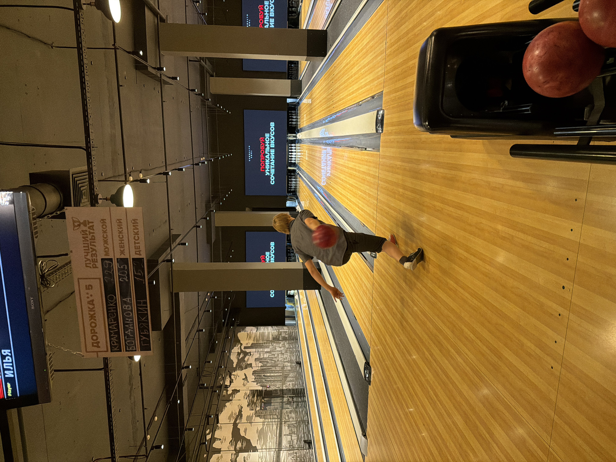 The image depicts a young person, viewed from behind, in the process of throwing a red bowling ball down a wooden lane. The person is wearing a gray t-shirt and dark shorts. In the background, several bowling lanes extend into the distance, lined with bowling pins at the far end. Large screens display graphics and text, one of which includes the text "ПОПРОБУЙ УНИКАЛЬНОЕ СОЧЕТАНИЕ ВКУСОВ" (Try a unique combination of flavors). To the left, a scoreboard displays names and scores, including "ДОРОЖКА: 5" (Lane: 5), "КРАМАРЕНКО 229 МУЖСКОЙ" (KRAMARENKO 229 MALE), "БОГДАНОВА 205 ЖЕНСКИЙ" (BOGDANOVA 205 FEMALE), and "ГУБЯКИН 157 ДЕТСКИЙ" (GUBYAKIN 157 CHILD). The environment appears to be an indoor bowling alley with overhead lighting and tall pillars. The city of Izhevsk, Russia, is not explicitly identifiable from the visual elements in the image; however, the text on the scoreboard and screen are in Russian, suggesting a Russian-speaking location.