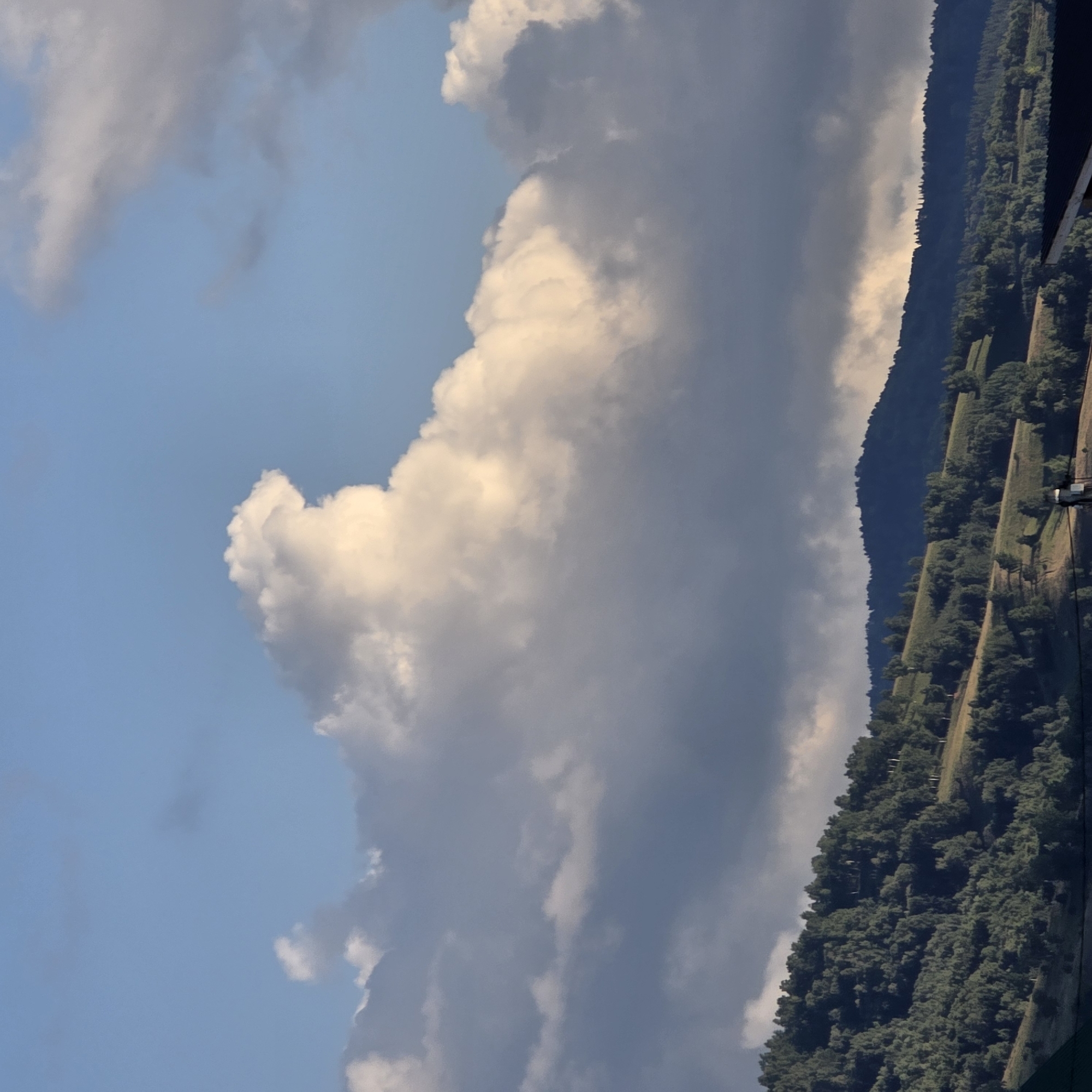 The image captures a scenic view of rolling hills covered in dense green trees and shrubs under a partly cloudy sky. The hills are characterized by a textured landscape with a mix of light and shadow, suggesting the sun is either rising or setting. Scattered within the vegetation are patches of lighter green, indicating cleared areas or different types of foliage. In the foreground, the dark silhouette of a rooftop is visible, partially obscuring the base of the hills. The sky is a blend of light blue and expansive white and gray clouds, with a particularly prominent, luminous white cloud formation in the upper center of the frame. This scene is typical of the natural beauty found in the vicinity of Belokurikha, Russia, a resort town known for its mountainous terrain and forested landscapes.