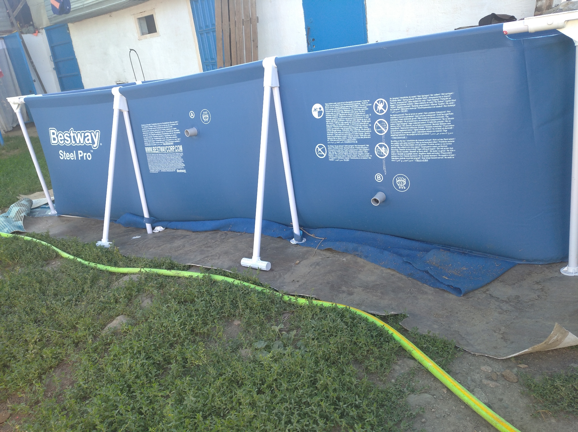 The image shows a blue Bestway Steel Pro above-ground swimming pool set up on a concrete pad next to a grassy area. A yellow garden hose snakes across the grass and near the edge of the pool. The pool is supported by white metal poles. In the background, there is a white building with blue doors and a small window. The scene appears to be in an outdoor setting. There are no people visible in the image. The location is not specified in the image and cannot be determined.