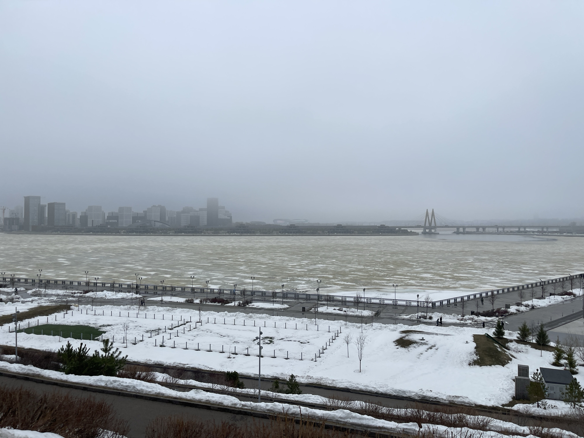 This image depicts a winter scene in Kazan, Russia, specifically showing a view of the Kazanka River and the cityscape beyond it, obscured by overcast skies. The river is largely frozen over with patches of open water visible. A long, concrete pier or promenade extends along the river's edge, lined with lampposts and railings.
In the foreground, there's a park covered in snow. Some patches of green grass are visible. There are structures with snow on them. A two-lane road cuts through the bottom of the image, and some brush and foliage are visible in the very bottom of the frame.
In the distance, the hazy skyline of Kazan is visible, including modern buildings and a suspension bridge across the Kazanka River. Several pedestrians are walking along the pier. The sky is uniformly gray.