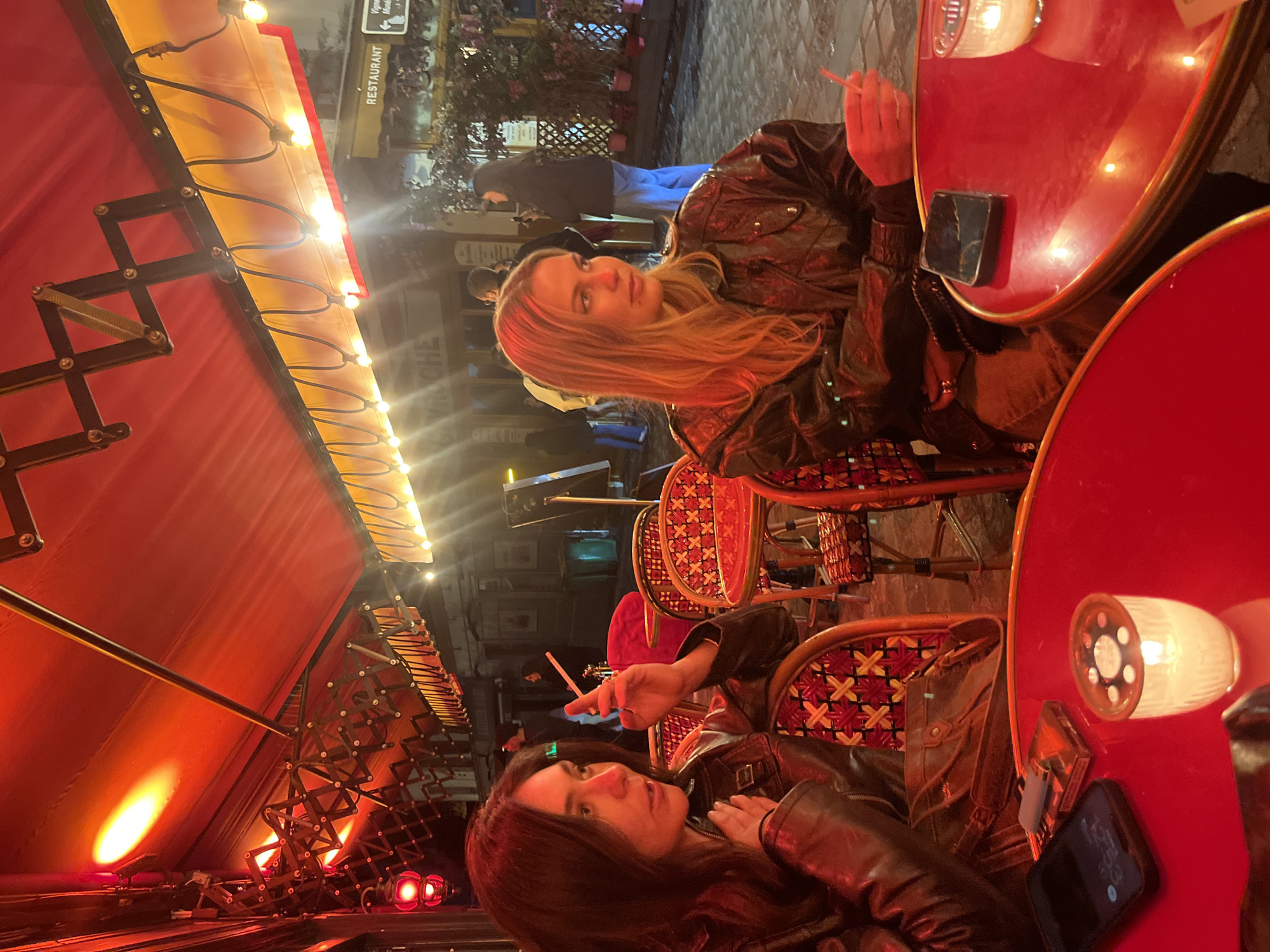 The image shows two women sitting at a red table outside a restaurant in Paris, France. Both women are wearing leather jackets and are holding cigarettes. The woman on the right has long blonde hair, while the woman on the left has long dark hair. The table has a phone and a cigarette pack on it. The restaurant has a red awning with lights and patterned chairs. In the background, people walk on the street. A sign reads "RESTAURANT".