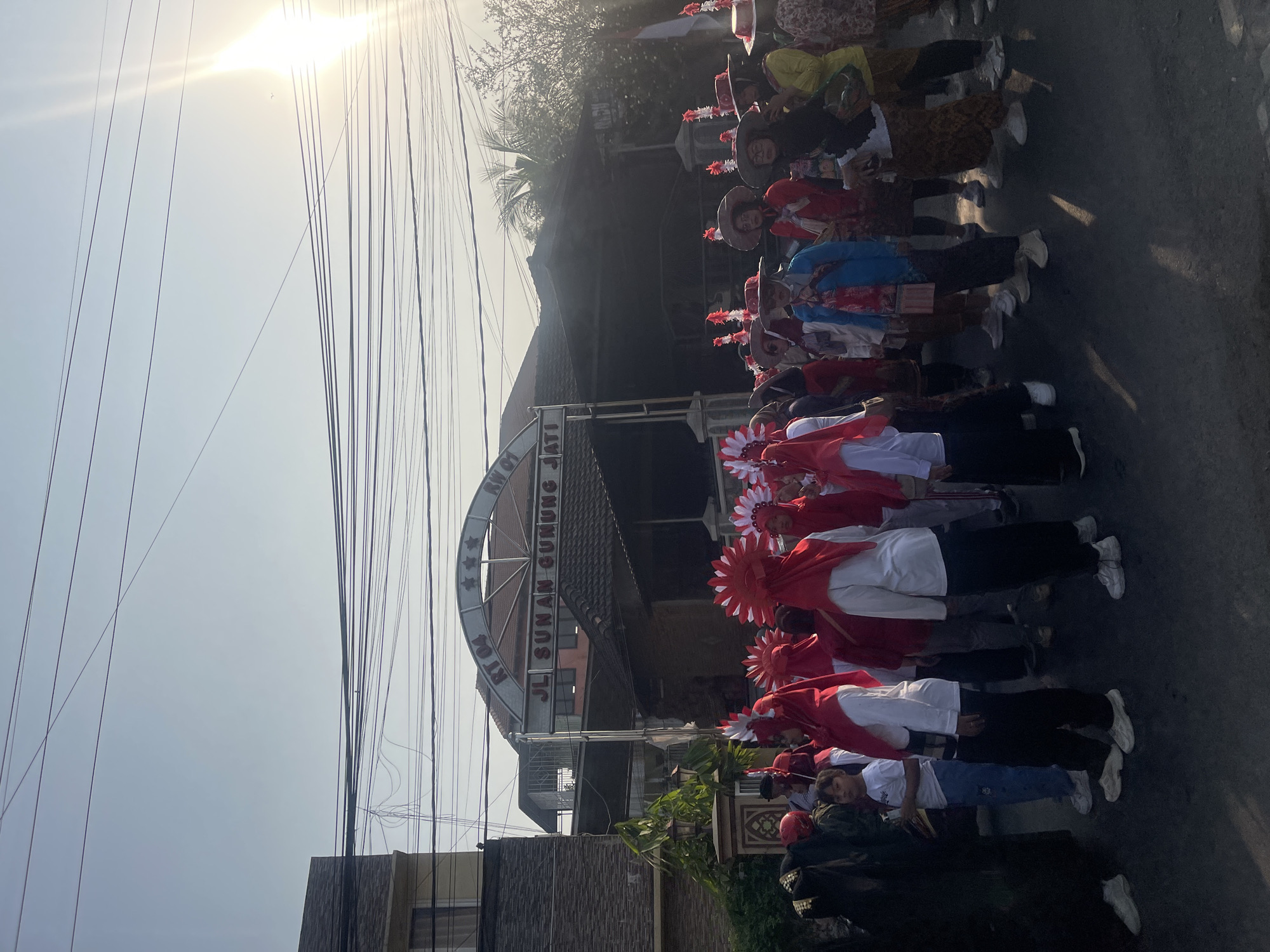 The image depicts a street scene in Sugih Waras, Indonesia, with a group of people participating in a parade or procession. The sun is shining brightly, casting long shadows on the asphalt road. In the background, an archway with the inscription "JL SUNAN GUNUNG JATI" is visible, indicating the location.
The participants are predominantly women, many of whom are wearing red headscarves and white tops, suggesting a uniform or costume for the event. Some are also wearing traditional Indonesian patterned skirts. Several individuals are adorned with elaborate red and white headdresses. In the foreground, a young boy in a white t-shirt and blue pants stands out. The group appears to be moving forward as a unit. Overhead, a dense network of power lines crisscrosses the sky, typical of urban or suburban Indonesian environments. The overall atmosphere suggests a festive or ceremonial occasion.