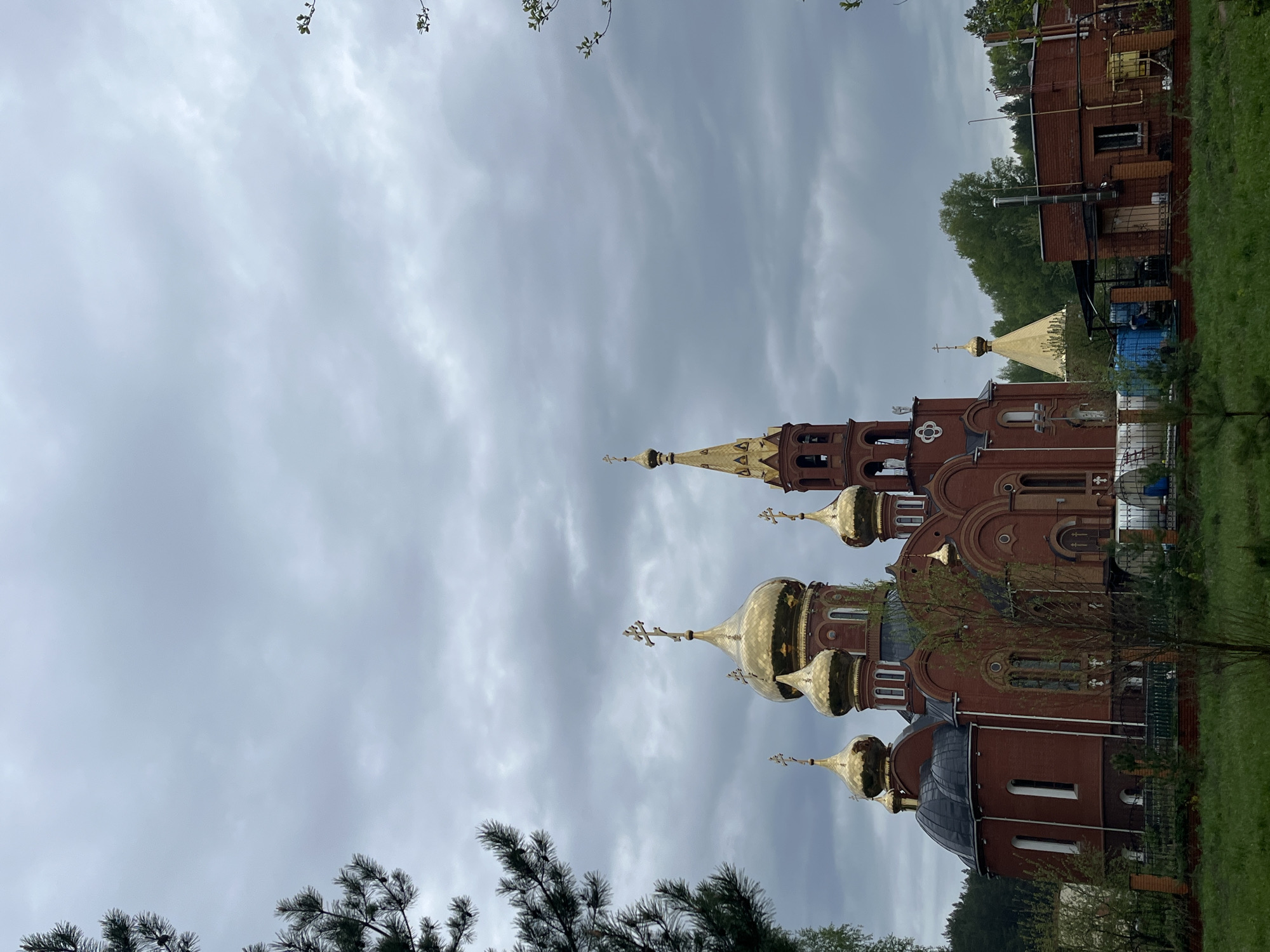 The image features a red brick Orthodox church in the city of Chaikovsky, Russia. The church has multiple golden domes with crosses on top. The sky is overcast. Trees are visible in the foreground and background. A brick building is located next to the church.