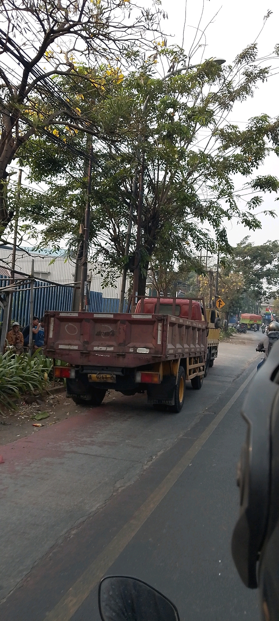 In Bekasi, Indonesia, a weathered, maroon-colored dump truck is parked on the side of a road. The truck bed is empty, showing signs of wear and tear. Behind the truck, a blue metal fence lines a property with a light-colored building visible beyond it. A few individuals are seated on a low wall near the fence, observing the surroundings. Lush green plants grow along the roadside and behind the fence. A bright yellow street sign with a black arrow is visible further down the road, indicating a turn. Several motorcycles are parked on the right side of the road, and a motorcyclist wearing a helmet is in motion, approaching the viewer. The road surface is paved and shows some cracking, with a yellow line marking the lane. Trees with sparse green leaves and some yellow blossoms are abundant, with power lines crisscrossing the sky above.