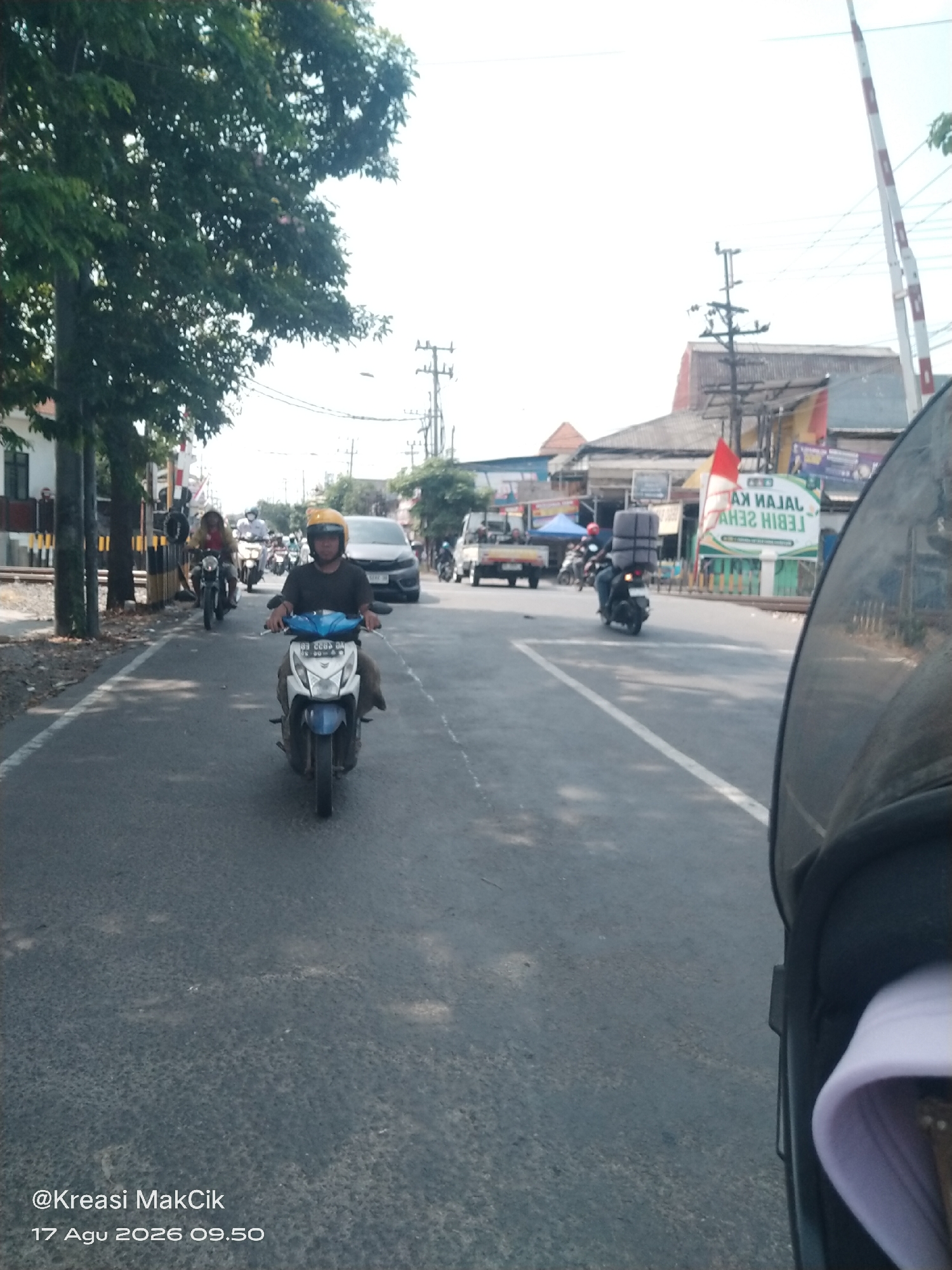 On a sunny day, a person wearing a yellow helmet is riding a blue and white motorcycle on a paved road in Kalijaten, Indonesia. The rider is wearing a dark t-shirt and light-colored pants. The motorcycle's license plate is visible. In the background, other motorcycles and a car are also present on the road. To the left of the road, there are trees and buildings. A railroad crossing signal is visible in the distance. A white sign with red lettering is also visible in the background. The bottom of the image contains text that reads "@Kreasi MakCik 17 Agu 2026 09.50".