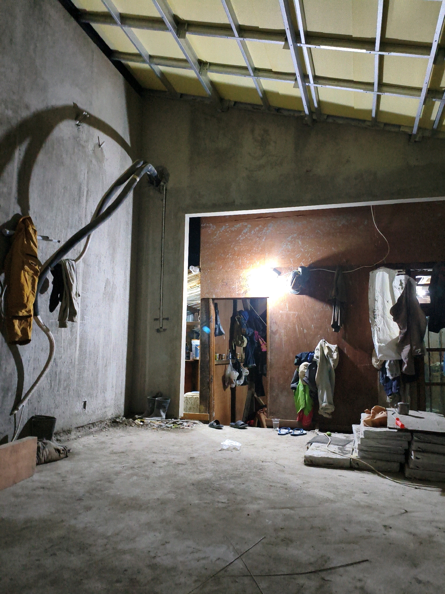The image depicts an unfinished interior space, likely a room under construction or renovation, located in the city of Bekasi, Indonesia. The walls are bare concrete, and the ceiling frame, made of metal supports, is visible with insulation panels above. Plumbing pipes run along the left wall, and several pieces of clothing are hanging from hooks on this wall. The floor is concrete and appears somewhat dusty, with some debris scattered around.
In the background, there is an open doorway leading to another area. The walls of this adjacent space are a reddish-brown color, and a bright light source illuminates the area, casting strong shadows on the wall. More clothing items are hanging in this doorway and on the reddish-brown wall. Several pairs of rubber boots and sandals are visible on the floor near the doorway, along with stacks of concrete slabs or tiles on the right side of the image. The overall impression is one of a utilitarian and work-in-progress environment.