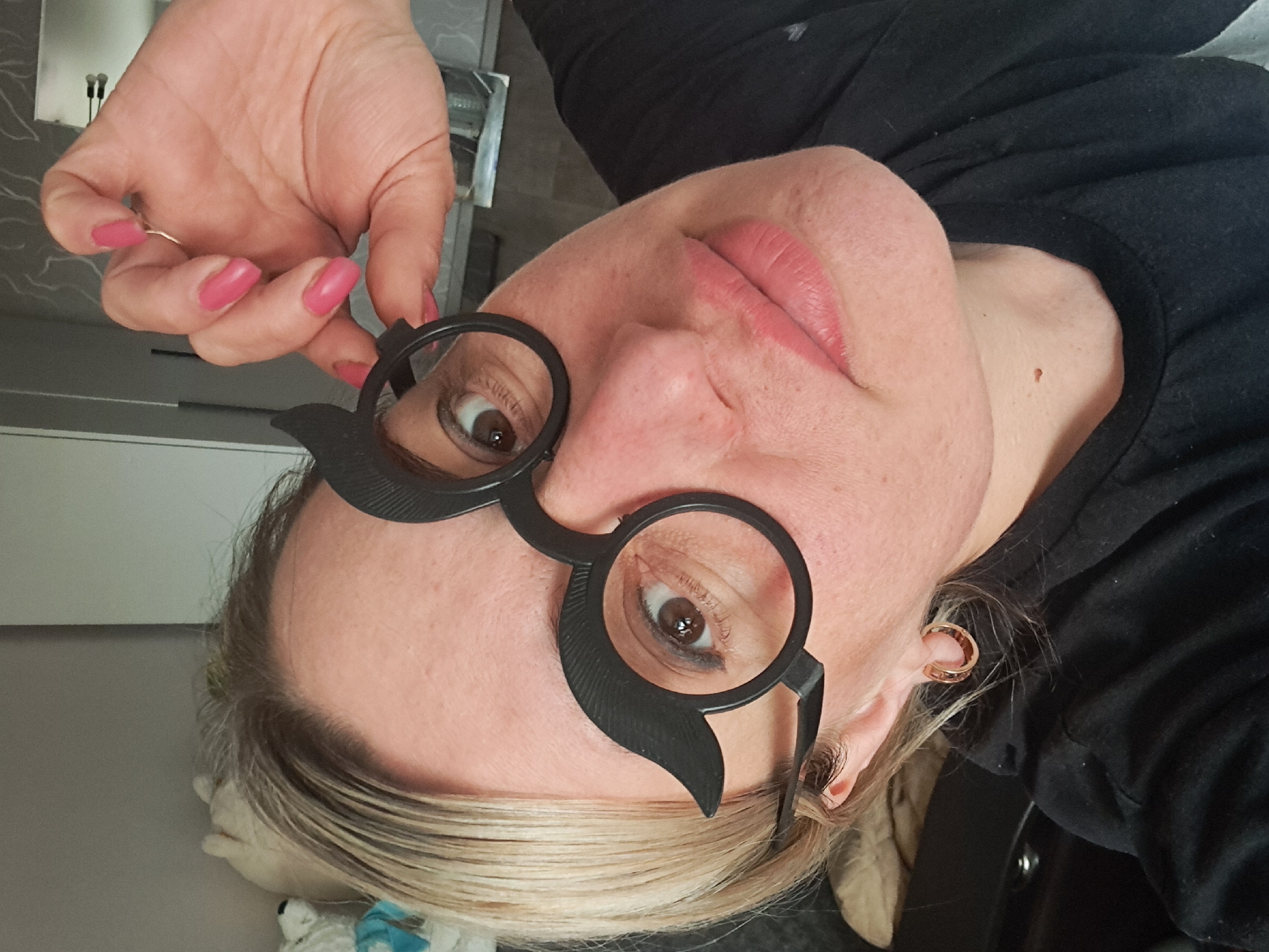 A woman with fair skin, blonde hair, and pink nail polish is holding up a pair of black, round novelty glasses with mustache shapes attached to the sides. She is wearing a black shirt and a gold hoop earring is visible in her ear. The location appears to be indoors. It is not possible to definitively determine the location as Elektrostal, Russia from this image.