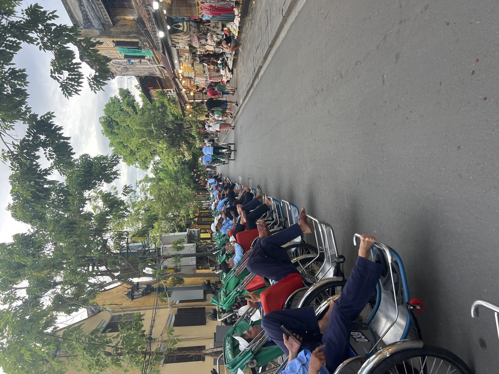 The image depicts a street scene in Hoi An, Vietnam, likely during the late afternoon or early evening, given the lighting. The focus is on a row of parked cyclos, a common mode of transportation in the city. Several men, presumably cyclo drivers, are seated in or near their vehicles, some of whom appear to be resting or using their mobile phones. The cyclos themselves are distinctive with their green canopies and red seating.
On the right side of the street, there is a row of shops or stalls, with their awnings and goods displayed, suggesting a bustling marketplace. Bags, clothing, and various trinkets are visible. Pedestrians are walking along the sidewalk and street, some browsing the shops. The architecture of the buildings lining the street is characteristic of Hoi An, with yellow-toned facades and traditional window designs. Lush green trees with dense foliage are scattered throughout the scene, providing shade and adding to the visual appeal. The sky is overcast, with a muted light. The overall atmosphere is one of a lively, yet relaxed, urban environment.
