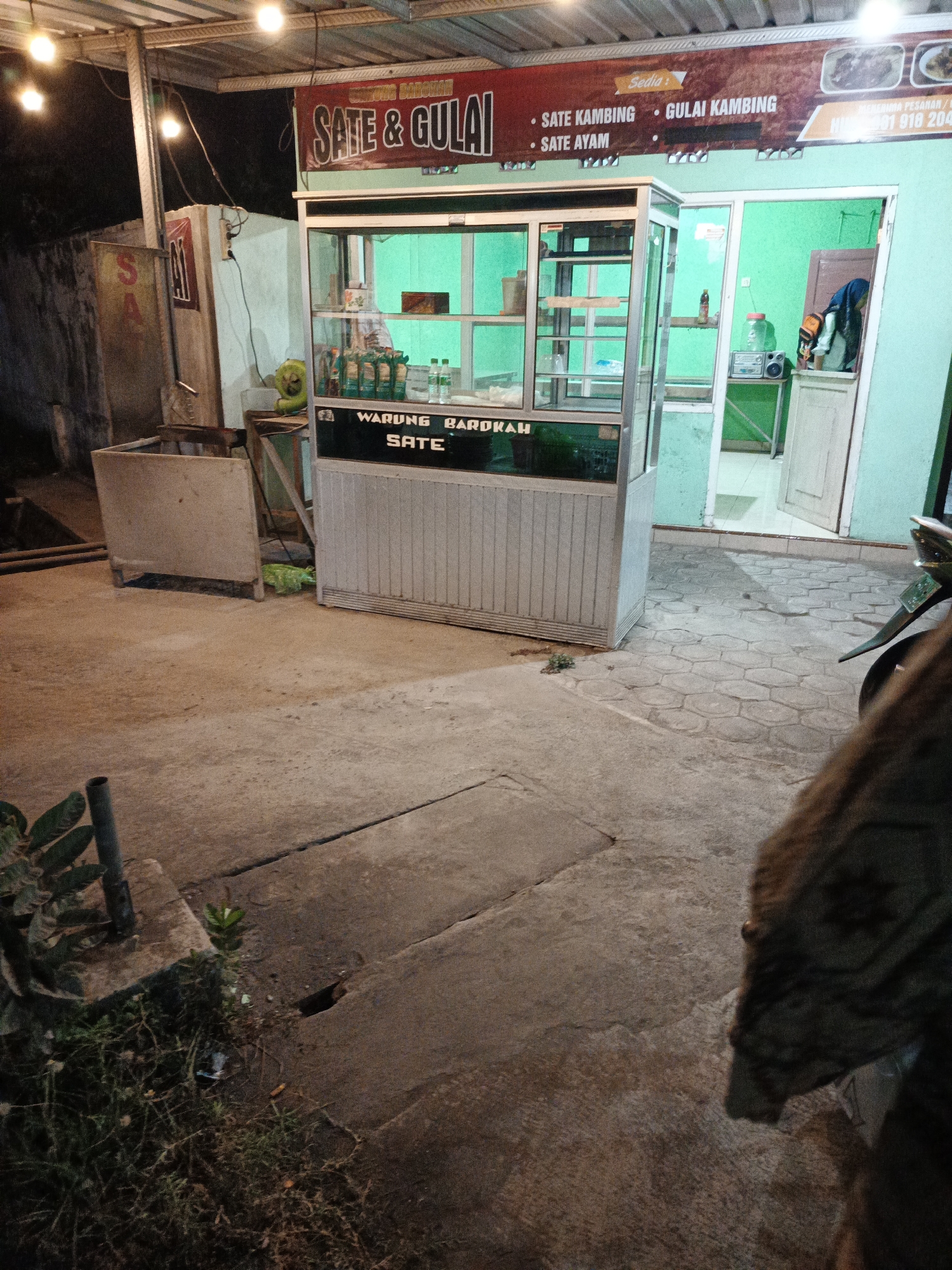 The image depicts a small food stall selling "Sate & Gulai" (satay and curry) in Sumbersari, Indonesia. The stall features a glass display case filled with various items, likely ingredients for the dishes. A prominent sign above the stall advertises "SATE KAMBING" (goat satay) and "SATE AYAM" (chicken satay), as well as "GULAI KAMBING" (goat curry). In the background, a green wall and an open doorway suggest the stall is attached to or adjacent to a building. The ground is paved with concrete and hexagonal tiles. A motorcycle is partially visible on the right side of the frame, indicating a common mode of transport in the area. The scene is lit by overhead lights, suggesting it is nighttime or dusk.