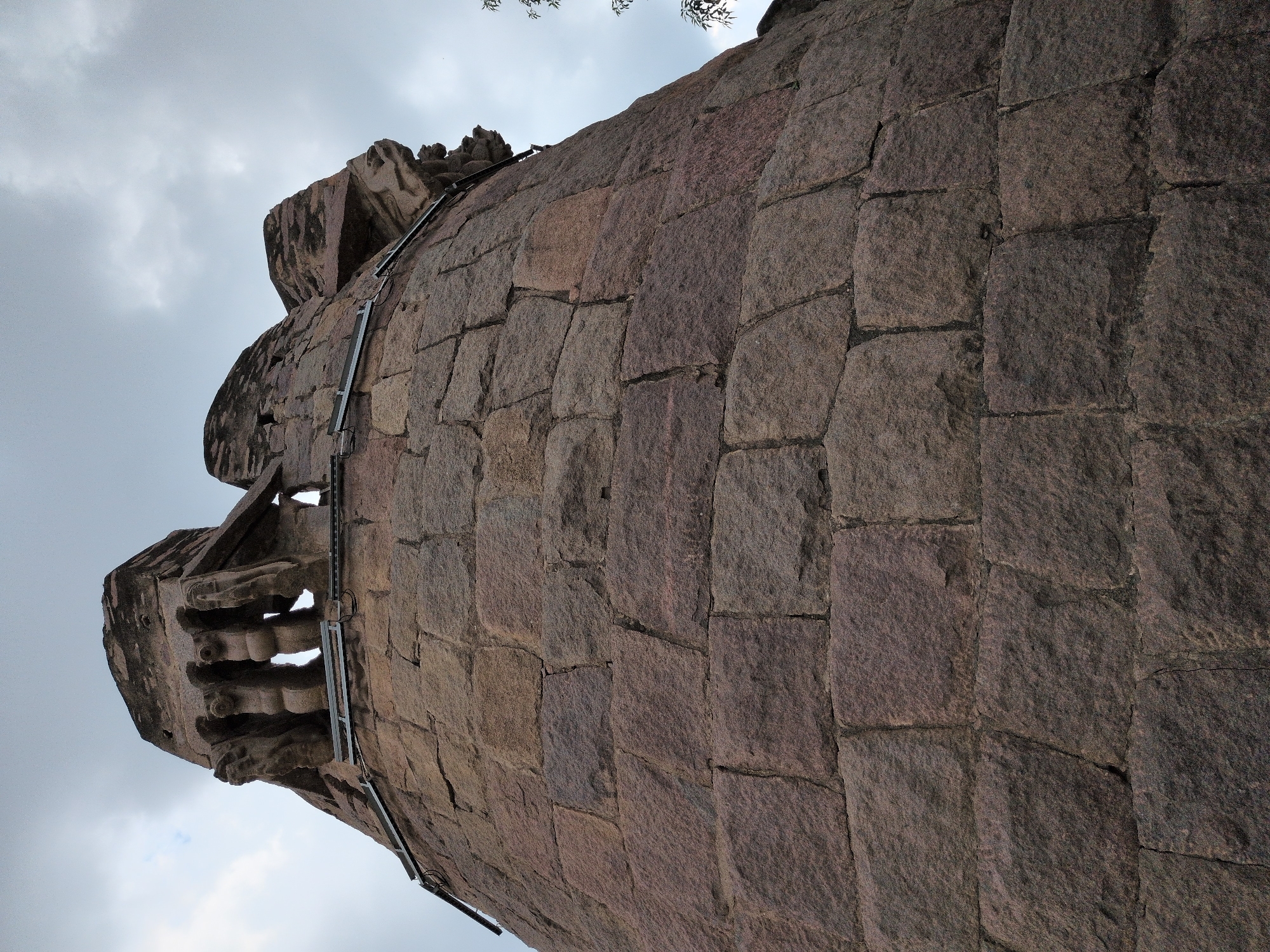 This image captures a low-angle perspective of a massive, circular stone bastion or tower, part of the historic Golconda Fort in Hyderabad, India. The structure is built from large, roughly hewn, dark reddish-brown granite blocks, exhibiting an ancient yet robust masonry style.
The upper section of the bastion features intricate stone carvings. On the visible left side, three prominent carved supports or corbels are discernible, depicting stylized lion-like or mythical 'Yali' figures with distinct heads, faces, and paw-like elements, appearing to brace an overhanging element of the battlements. To the right, another similar carved section is partially visible. Directly below these carvings, horizontally mounted metallic rails or bars with associated wiring are affixed to the stone, indicative of modern lighting installations or structural enhancements. The sky above is overcast with diffused grey clouds. A sparse cluster of green leaves from a tree is visible on the extreme right edge of the frame. The low-angle vantage point emphasizes the towering height and imposing scale of the ancient fortification.