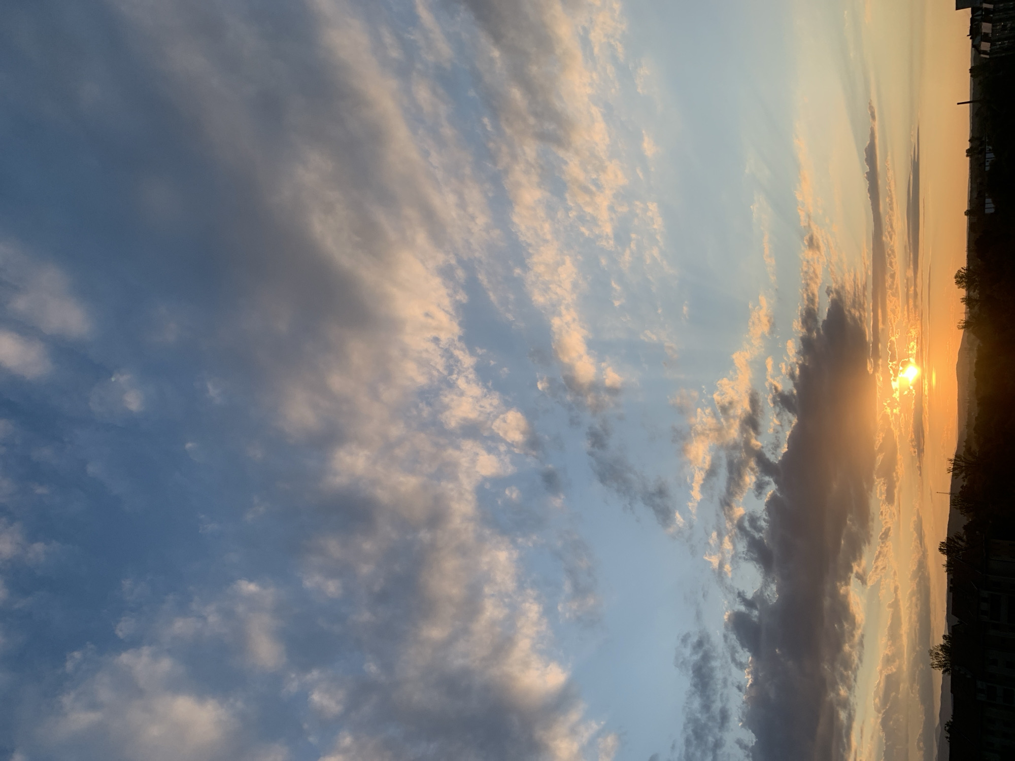 The image shows a sunset over the city of Kolomyia, Ukraine. The sun is setting behind a range of hills in the distance, casting a warm glow over the landscape. The sky is filled with a mix of clouds, some of which are illuminated by the setting sun with shades of orange and pink. In the foreground, the silhouettes of buildings and trees are visible against the colorful sky. The overall impression is one of a peaceful and serene evening.