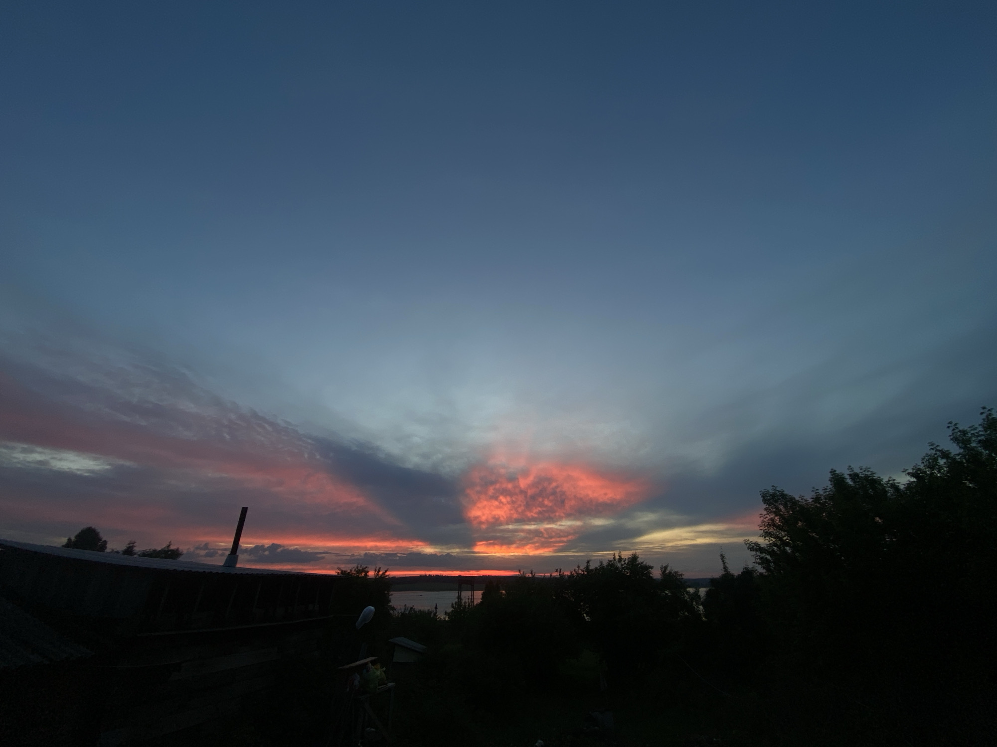 The image captures a vibrant sunset or sunrise over a natural landscape in Smolino, Russia. The sky dominates the upper two-thirds of the frame, transitioning from a deep, muted blue-grey at the top to striking reds, oranges, and pinks closer to the horizon. Wispy and denser clouds are illuminated by the low sun, creating dramatic, fan-like rays of color that spread across the sky.
At the horizon, a calm body of water, likely a lake or river, is visible as a thin strip of lighter blue and grey, reflecting some of the sky's soft glow. A faint, distant shoreline can be discerned on the opposite side.
The foreground is composed almost entirely of dark silhouettes. On the left, the dark roofline of a building or shed is prominent, with a tall, dark pipe-like structure (possibly a chimney or vent) rising from it. Below the roofline, indistinct shapes suggest other structures or debris. To the right and across the bottom of the frame, dense masses of trees and foliage create a dark, undulating silhouette, obscuring any ground details and framing the colorful sky.
The time of day is clearly either dusk or dawn, given the intense and varied sky colors. The weather appears calm, with enough clouds to enhance the visual spectacle. There is no visible text in the image. The overall impression is one of serene beauty and natural grandeur.