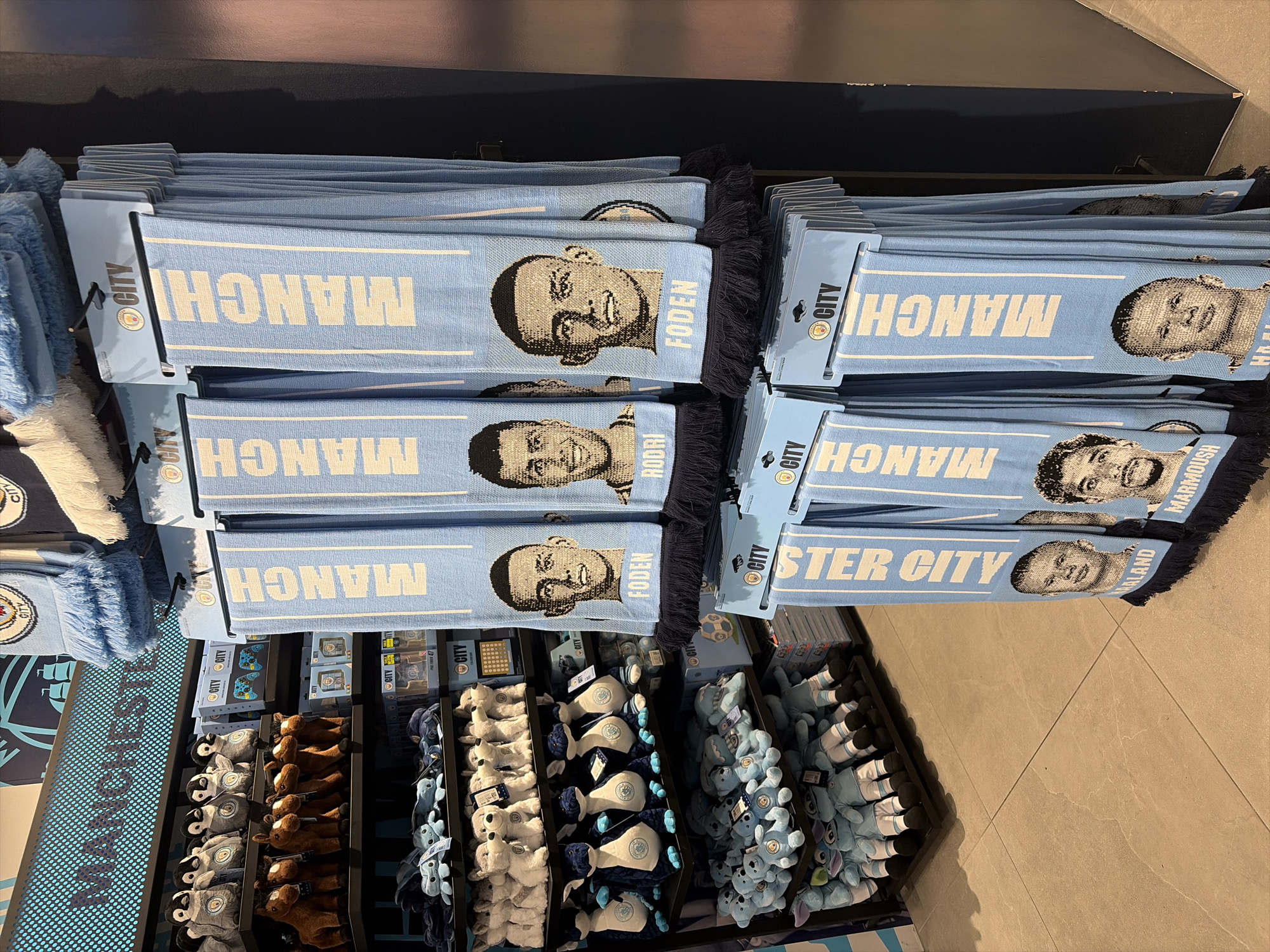 The image shows a display of Manchester City football club merchandise. The display features stacks of light blue scarves with the word "MANCH" printed in white and the faces of football players Rodri, Foden, Haaland and Marmoush. Below the scarves is an assortment of plush toys of various Manchester City team mascots and team colors. The environment appears to be a store, likely in the city of Manchester, United Kingdom, based on the club affiliation.