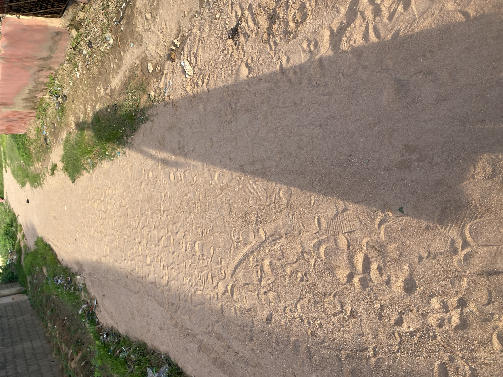 The image depicts a sandy path in Jalingo, Nigeria, with numerous footprints visible on the surface. To the left of the path is a wall constructed of grey bricks, bordered by sparse green grass and some scattered litter. To the right, a building with a reddish-pink wall is visible, also with some debris on the ground. A long shadow of a person, cast by the sun, stretches across the path, indicating the presence of an individual or individuals who have traversed this route. The environment appears to be an outdoor, unpaved area within a settlement.