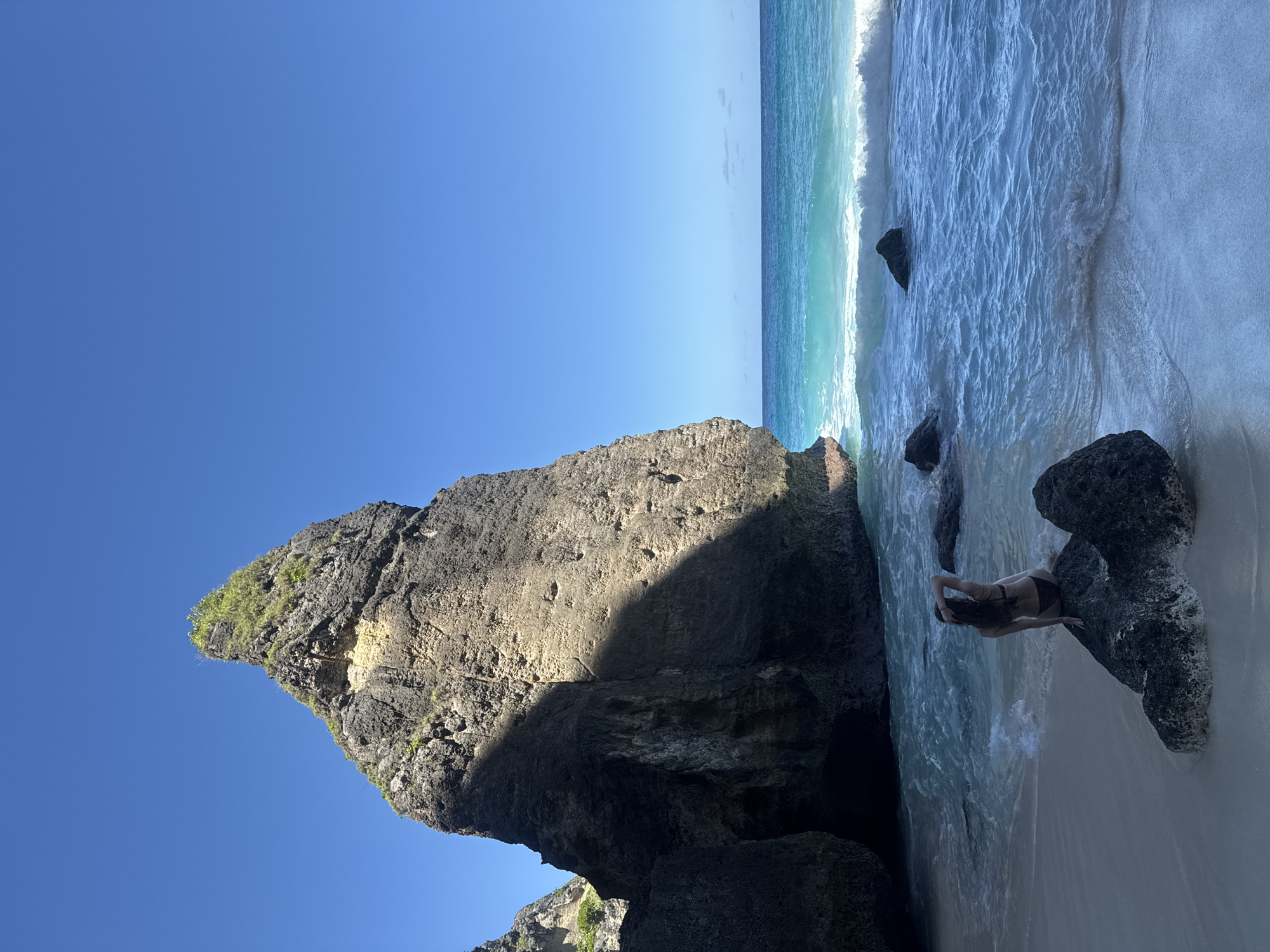 The image shows a rocky beach on a sunny day. The location is Nusa Penida, Indonesia, as suggested by the scenery and the presence of a large, distinctive rock formation. The sky is a clear, deep blue, contrasting with the turquoise water. A significant portion of the image is occupied by a large, dark-colored rock formation. Part of the rock is in sunlight, and part is in shadow. Vegetation is visible on the top of the rock formation. The ocean water is clear and turquoise, with visible waves. A person in a bathing suit is present in the shallow water near the shore. The person is standing among the rocks.