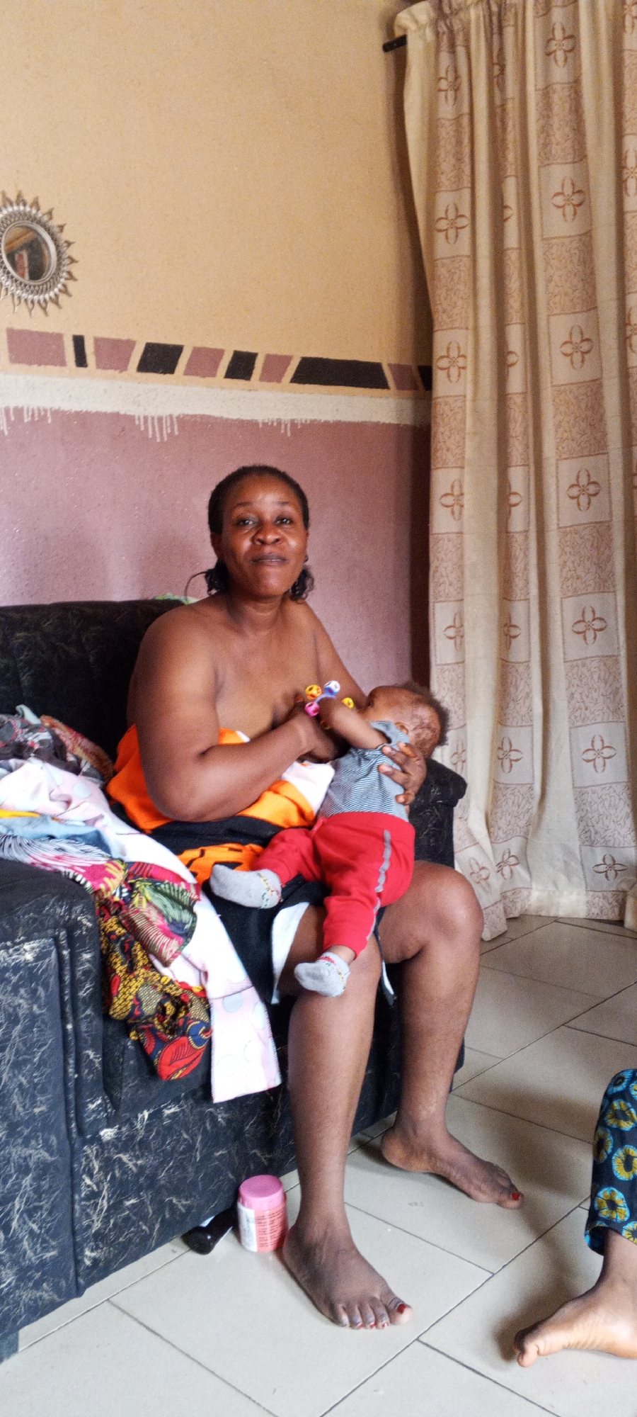 A woman is seated on a dark-colored sofa, breastfeeding a baby. The woman is dark-skinned and has short, dark hair. She is bare-chested with an orange garment draped over her shoulder. The baby, dressed in a gray and white striped shirt and red pants, is being held close to the woman's chest. A colorful pile of clothes is on the sofa next to the woman. The room has beige walls with a decorative band near the ceiling. A round mirror hangs on the wall. A beige curtain with a floral pattern is visible to the right of the sofa. On the floor, near the woman's feet, are a pink container and another person's feet wearing blue patterned pants. The scene appears to be indoors, possibly in a home located in Sabon Tasha, Nigeria, given the prompt.