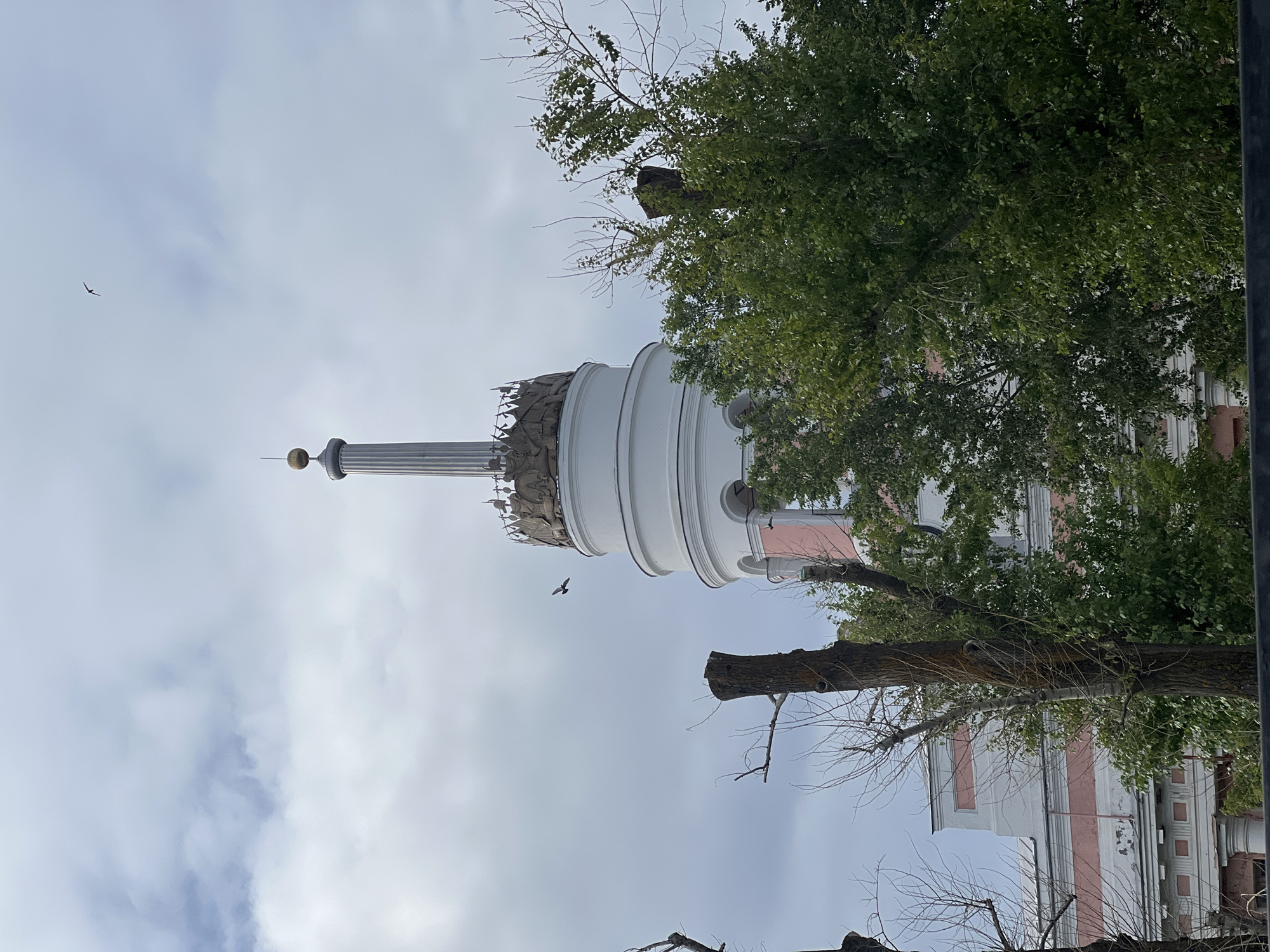The image displays a prominent, multi-story building in Izhevsk, Russia, primarily focusing on its distinctive tower against an overcast sky. The tower, situated centrally in the frame, features a white cylindrical base with visible arched openings. Above this, a bronze-toned frieze depicting a continuous band of relief figures, appearing humanoid and in historical or allegorical poses, encircles the structure. Surmounting this section is a tall, slender, fluted column or spire, topped with a golden spherical finial from which a thin spike extends upwards.
Portions of the building visible below the tower feature a facade with horizontal pink stripes contrasting with white or light grey sections, punctuated by architectural cornices and pilasters. The sky is uniformly light grey, indicating overcast conditions, with visible cloud formations.
In the foreground and midground, several trees are present. Some trees display dense, vibrant green foliage, while others are severely pruned, appearing as dark brown trunks and stumps with minimal branching and an absence of leaves, suggesting recent trimming or dormancy. A prominent large, dark brown tree stump is located in the mid-left.
Two birds are observed in flight against the sky. A smaller, dark avian silhouette is visible in the upper right quadrant, while a second, larger dark bird flies closer to the tower, positioned to the left of its central column. The perspective is from a low vantage point, looking upwards towards the architectural structure and the sky.
