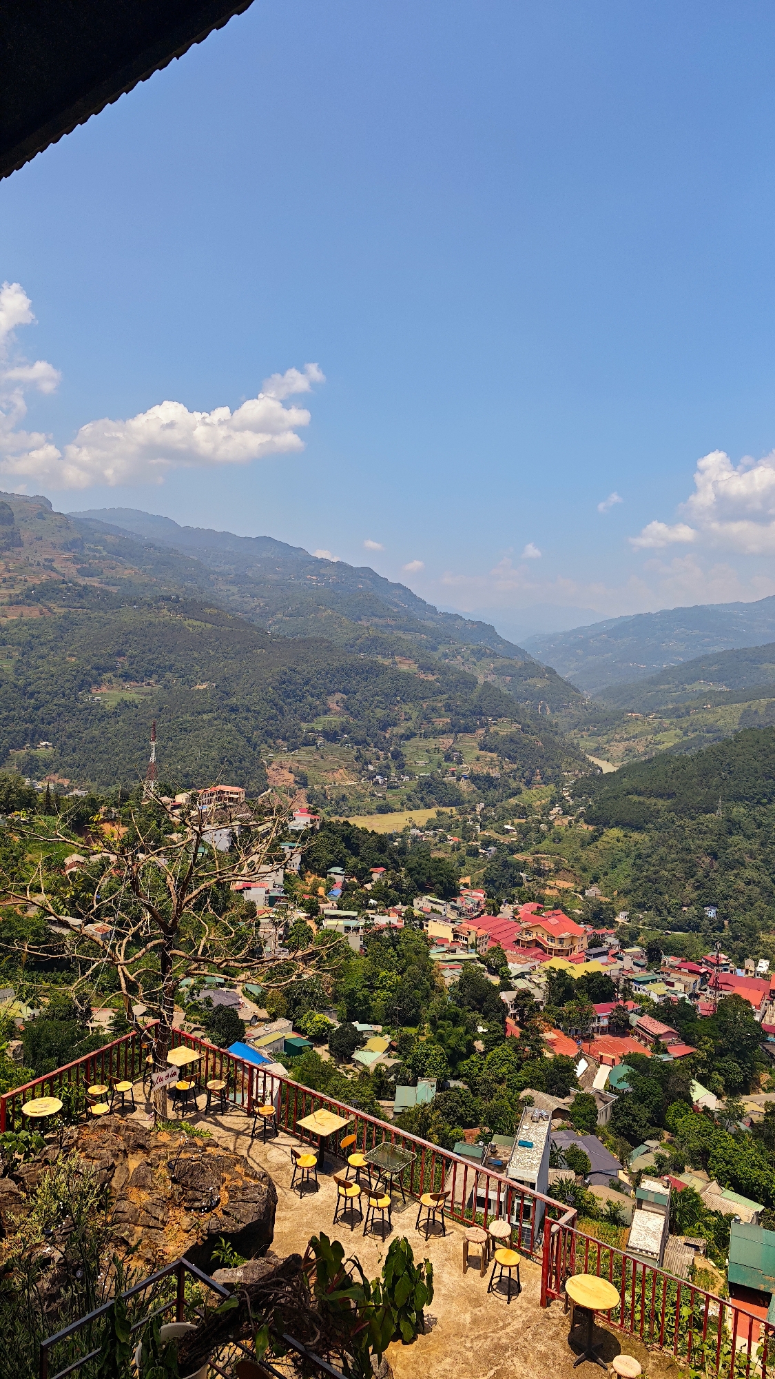 An elevated, high-angle shot captures a broad landscape in Cốc Pài, Vietnam, featuring mountainous terrain, a valley, and a town. The sky is bright blue with scattered white clouds.
In the foreground, an outdoor patio area is visible with reddish-orange railings. Several small, round tables with yellow stools are arranged on a concrete surface. Beyond the patio, a dense area of green trees and buildings with red roofs creates a central focal point.
In the midground, the town extends towards a valley flanked by mountains covered in dense vegetation. A tall communications tower is visible among the buildings. The background is filled with overlapping mountain ranges receding into the distance, contributing to the image's depth.