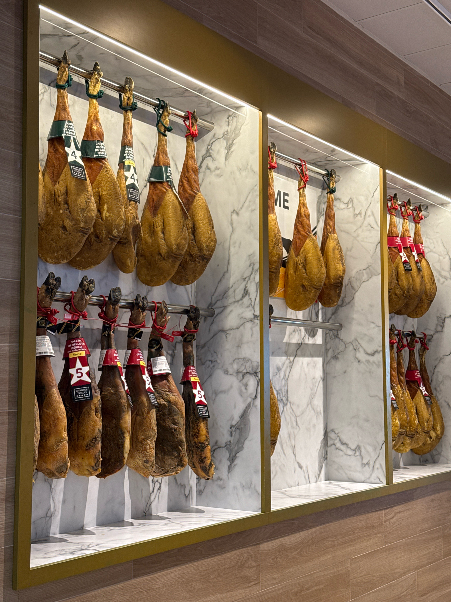 The image shows a display of cured hams hanging from metal rods within a store. The hams are arranged in rows, with some in the foreground and others further back. Each ham appears to have a label attached to it. The background consists of marble-patterned walls and a wooden floor. The lighting is bright and appears to come from overhead fixtures. There are no people visible in the image. The display is designed to showcase the hams in a clean and organized manner, suggesting a high-end food establishment. The overall impression is one of a specialty food shop or deli. Given the context of a previous query related to Sant Josep de sa Talaia, Spain, it is plausible that this establishment is located in that city.