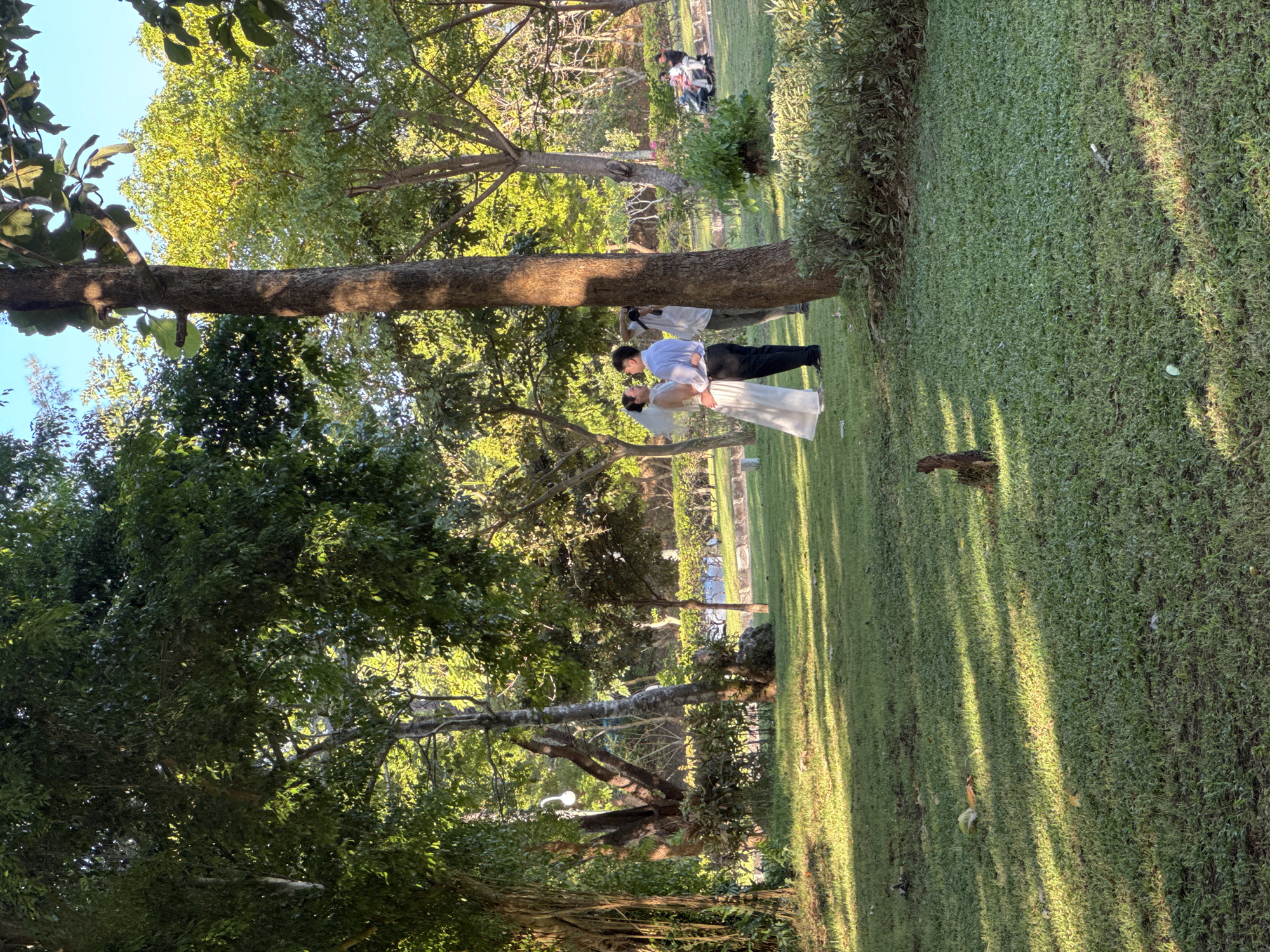 The image shows a couple, likely a bride and groom, in a park setting in Kuta Selatan, Indonesia. They are standing close, appearing to kiss or embrace. The groom is wearing a light-colored shirt and dark pants, while the bride is in a white dress. A person, possibly a photographer, is standing behind them. In the background, there are other people, trees, and well-maintained grass. The light suggests it is daytime, with the sun casting shadows across the lawn.