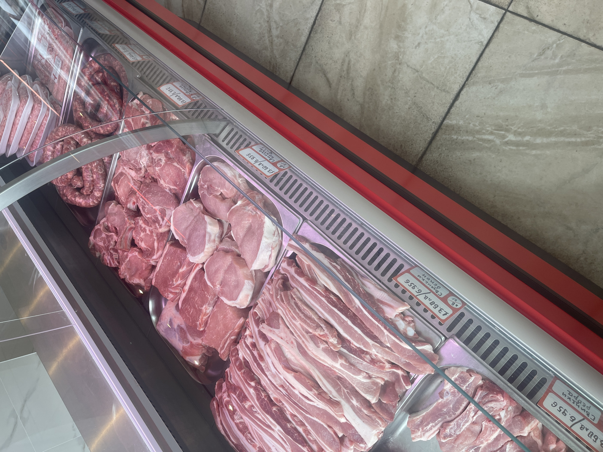 A close-up, high-angle shot captures a display case filled with raw meat, likely in a butcher shop or grocery store. The meat is arranged in trays, with various cuts visible, including what appears to be pork chops, ribs, and possibly some sausages in coiled form. Several price tags are visible, with pricing in Euros and Cyrillic script, indicating the location is likely in a European country where Cyrillic is used. The display case itself has a metallic finish and red trim, with a glass front reflecting light. The flooring beneath the display is tiled in a neutral, stone-like pattern. There are no people visible in the frame. The overall impression is one of fresh, raw meat neatly presented for sale.