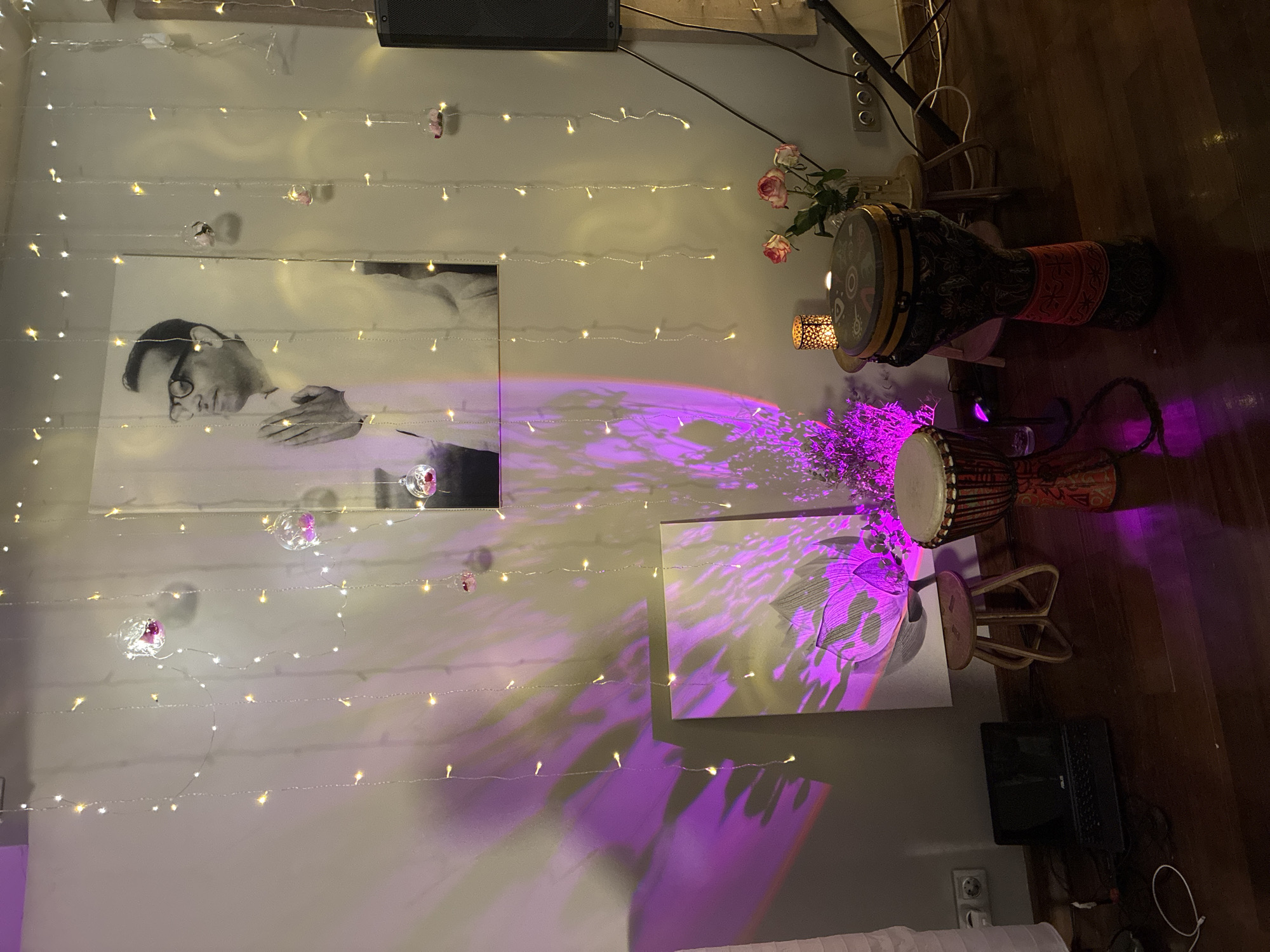The image presents an indoor scene, likely a room, characterized by a meditative or ceremonial ambiance, decorated with various elements. No individuals are present in the photograph.
The background wall is light-colored, possibly off-white or light gray, and is adorned with multiple vertical strands of warm white LED string lights. Interspersed among these lights are several small, clear spherical glass ornaments, each containing pink rose petals or similar small floral elements, hanging at various heights. Dominating the upper wall, slightly left of center, is a large, framed black and white portrait of a man wearing glasses, depicted with his hands pressed together in a prayer or greeting gesture (Anjali Mudra).
Below the portrait and to the left, a rectangular artwork featuring a lotus flower in muted tones leans against the wall. This artwork and the surrounding wall are bathed in a dynamic, wavy pattern of bright purple light projected from an unseen source to the left.
The room features dark wooden flooring. On the floor to the far left, a black laptop with a visible Asus logo is open, next to a white coiled cable and a double electrical outlet consistent with Type F/C standards found in Russia and Europe. Another double electrical outlet of the same type is visible on the wall to the right.
Two djembe drums are positioned on the floor: a smaller one with a red and patterned body to the left, and a larger one with a dark, intricate tribal pattern (including red accents) and a patterned drumhead to the right. A small, light-colored wooden stool with curved legs is placed near the smaller djembe. A second similar wooden stool to the right supports a clear glass vase containing a bouquet of wilting pink roses and two decorative candle holders, one a metal lantern with cutouts and the other a small dish with a lit candle.
To the far right, a large, black speaker enclosure is partially visible, next to a black stand, possibly for a microphone or other audio equipment. Dark cables run across the floor connecting these elements. The overall environment suggests a space prepared for a spiritual gathering, meditation session, or musical performance, with an emphasis on soft lighting and decorative elements. The electrical outlets are consistent with standards used throughout Russia and many parts of Europe, allowing for the possibility of the location being in Moscow, Russia.