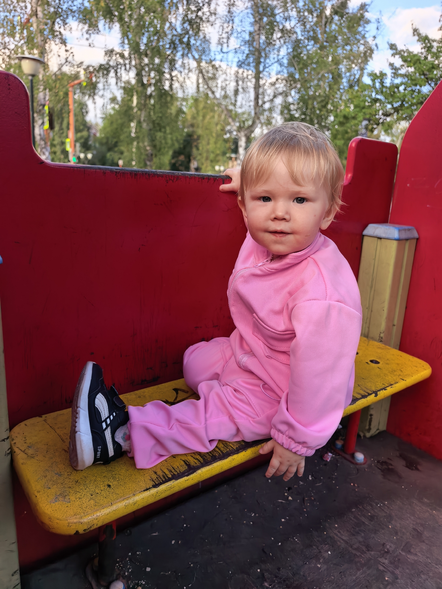 A young child with blonde hair, wearing a pink tracksuit and black and white sneakers, is sitting on a yellow bench attached to a red play structure. The child is looking towards the camera with a neutral expression. The background shows trees and a cloudy sky, suggesting an outdoor setting, likely a playground. The bench appears weathered and is set against a dark ground surface.