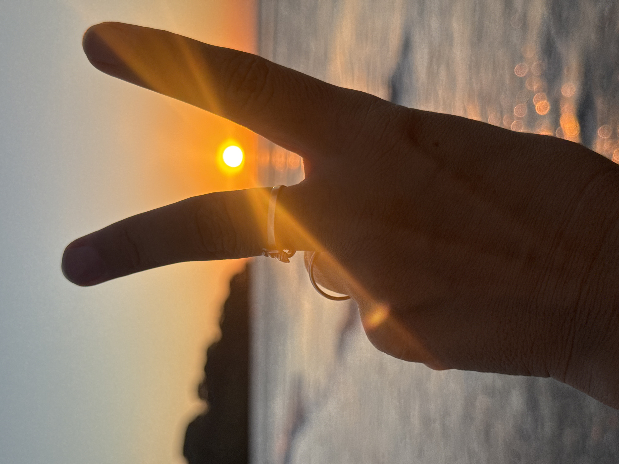 The image displays a human hand, silhouetted against a vibrant sunset or sunrise, forming a peace sign with the index and middle fingers extended. The sun, a small and intense yellow-orange circle, is precisely positioned between these two fingers, creating an optical illusion. A simple band ring is visible on the ring finger, illuminated by the strong backlight.
The scene is set over a body of water, likely the ocean, with shimmering light reflections (bokeh) visible on its surface, indicating the sun's low angle and the movement of the water. A hazy silhouette of distant land or an island can be faintly seen on the left horizon, adding depth to the background. Intense lens flare radiates from the sun, casting warm light streaks across the fingers and the upper right portion of the frame. The time of day is clearly either dawn or dusk, characterized by the warm, golden light and the sun's position. The overall color palette features warm oranges and yellows against darker tones of the silhouetted hand and landscape. This picturesque moment is captured in a coastal setting, specifically Kuta Selatan, Indonesia. There is no visible text in the media file.