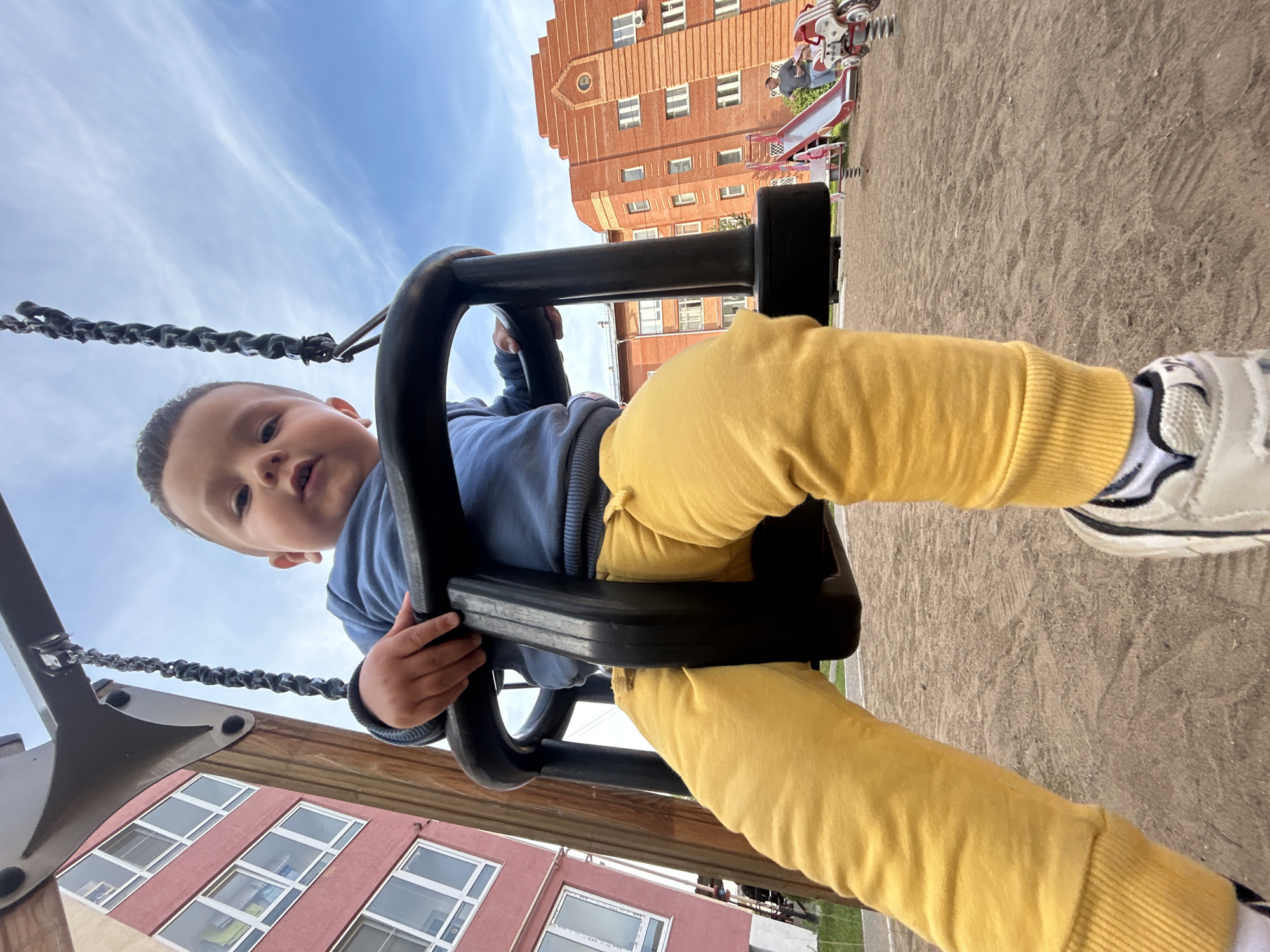 The image shows a young child on a swing in an outdoor playground. The child is wearing a blue long-sleeved shirt and yellow pants. They are holding onto the black swing seat with their right hand. A portion of their left leg and foot, wearing a white and black sneaker, is also visible. The background features a sandy ground, a red brick building with multiple windows, and a blue sky with some white clouds. A playground slide and other playground equipment are visible in the distance. The perspective is from the child's point of view looking upwards. The scene appears to be set during the daytime under a partly cloudy sky. The image conveys a sense of childhood fun and outdoor play. No visible text is present in the image.