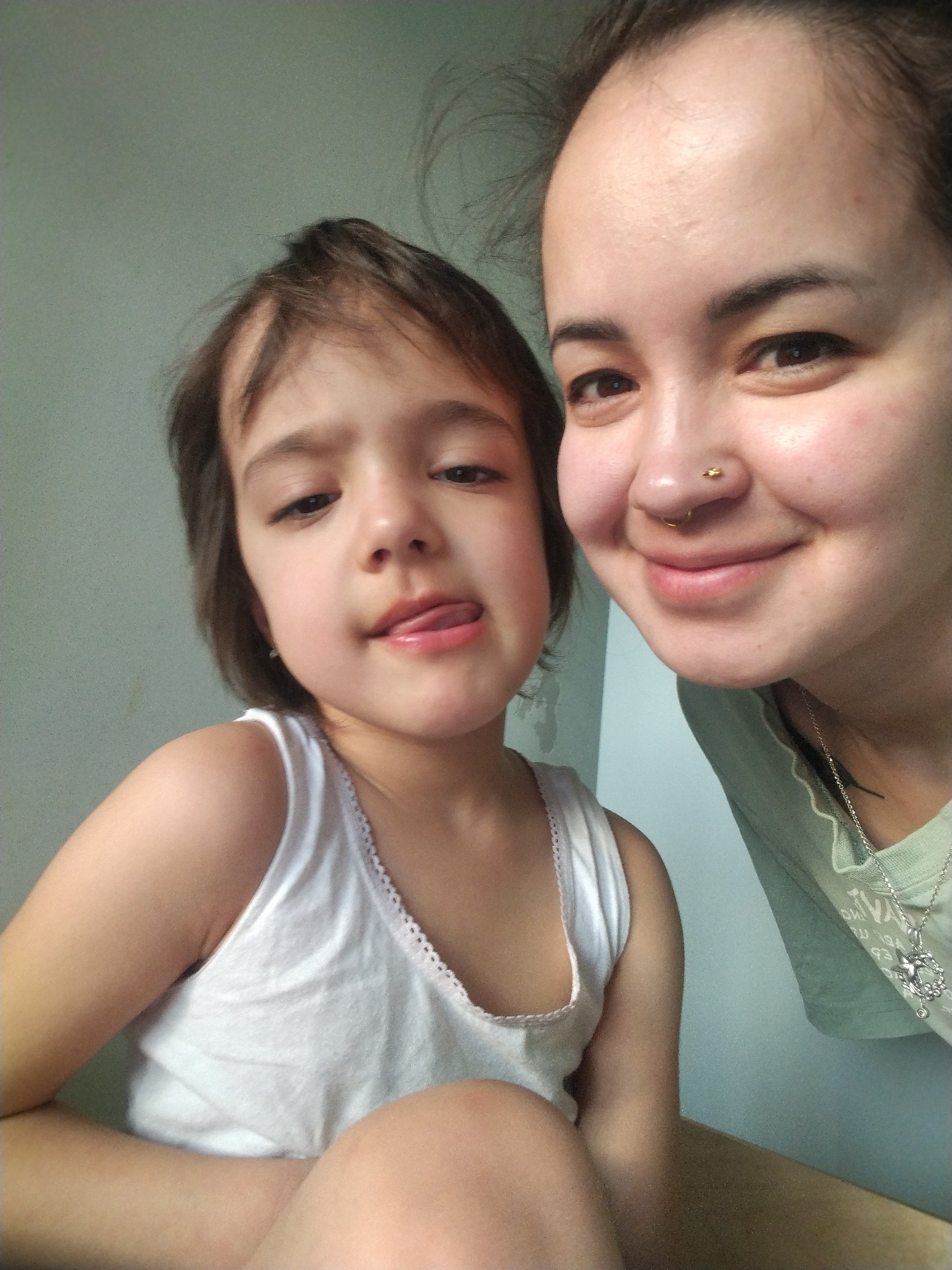 A selfie shows a woman and a child in a close indoor setting. The child, wearing a white tank top, is in the foreground, sticking her tongue out slightly. Behind her, a woman with a nose piercing smiles. The wall is a pale neutral color. A sliver of a light-colored surface is visible at the bottom of the frame. The location within Noviy, Russia, cannot be ascertained from the image.