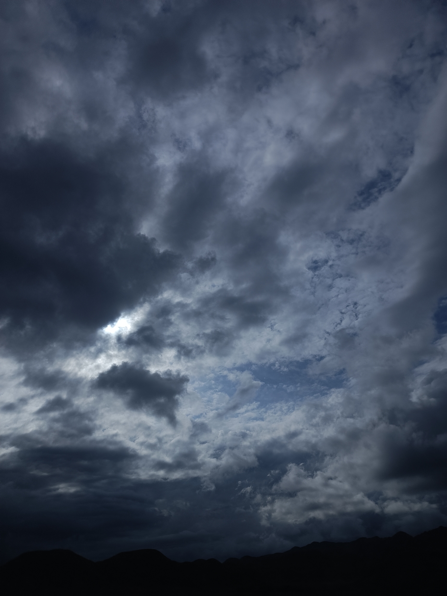A dramatic sky filled with dark, heavy clouds is the dominant feature of this image, with the silhouette of mountains visible at the bottom. The clouds are a mix of deep grays and blues, with a small patch of lighter sky and sunlight breaking through in the upper left quadrant. The overall mood is somber and foreboding, suggesting an approaching storm or the aftermath of one. The mountains appear to be in the distance, their peaks forming a jagged line against the turbulent sky. The location is identified as Sasyk-Bulak, Kyrgyzstan. There are no people or specific objects visible in the scene; it is purely a landscape focused on the atmospheric conditions.