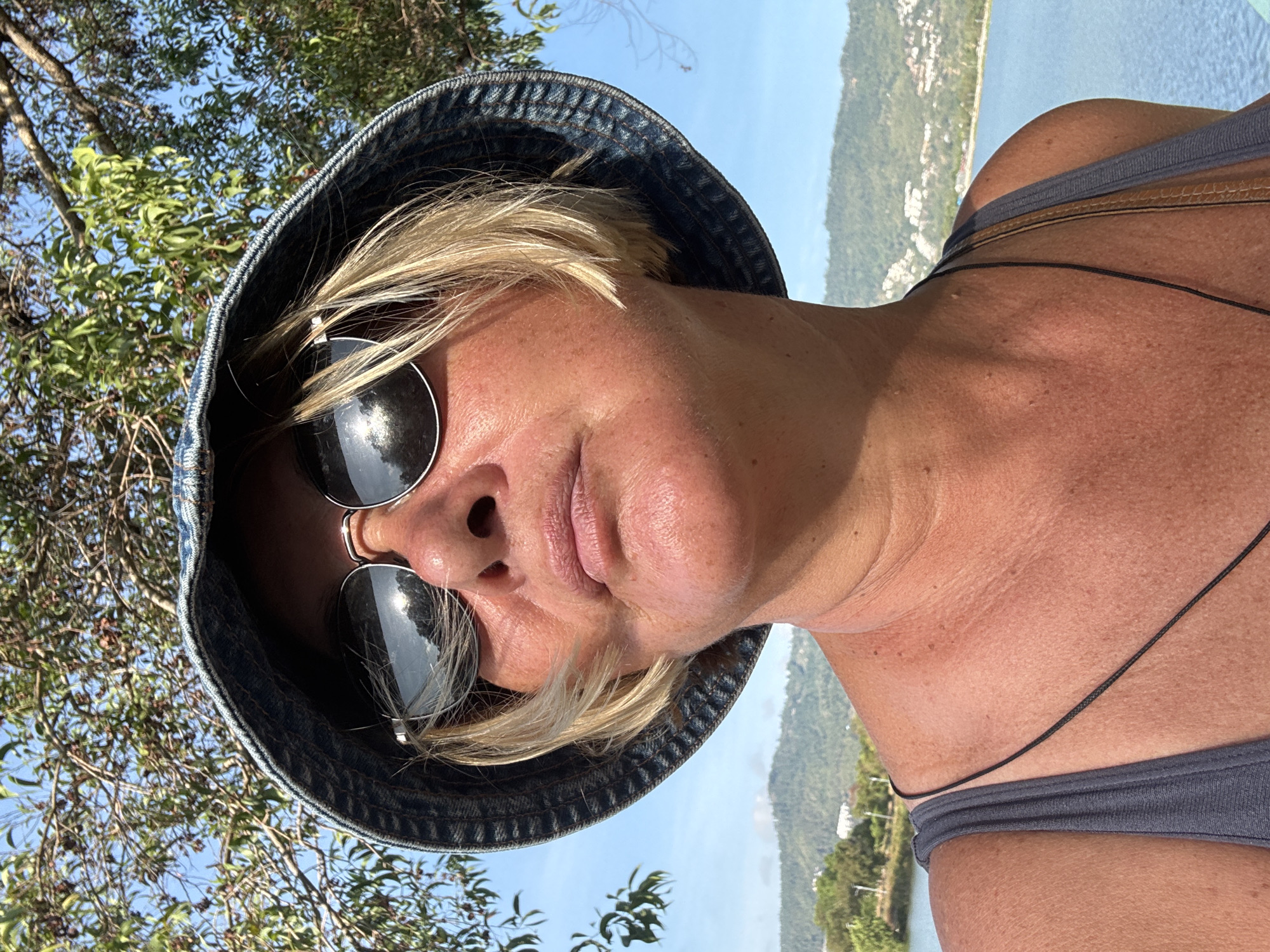 A close-up outdoor portrait features a woman with short, blonde hair, wearing a dark denim bucket hat and large, round, reflective sunglasses. Her face is partially visible, tilted slightly, with a subtle smile on her lips, suggesting a relaxed mood. Her skin appears tanned, indicating time spent outdoors. She is wearing a grey top or swimwear, with a thin black cord visible around her neck/shoulder.
The background reveals a sunny, clear day. Lush green trees and foliage are prominent, with bright blue skies above. Beyond the immediate greenery, a vast expanse of calm blue water, likely the sea or a bay, stretches into the distance. On the far shore, green hills are visible, dotted with scattered buildings, indicating a coastal landscape. The setting aligns with the provided context of บ้านบางรักษ์, Thailand, showcasing its natural beauty and sunny weather. The strong sunlight creates clear shadows, highlighting the brightness of the day. No text is visible in the image.