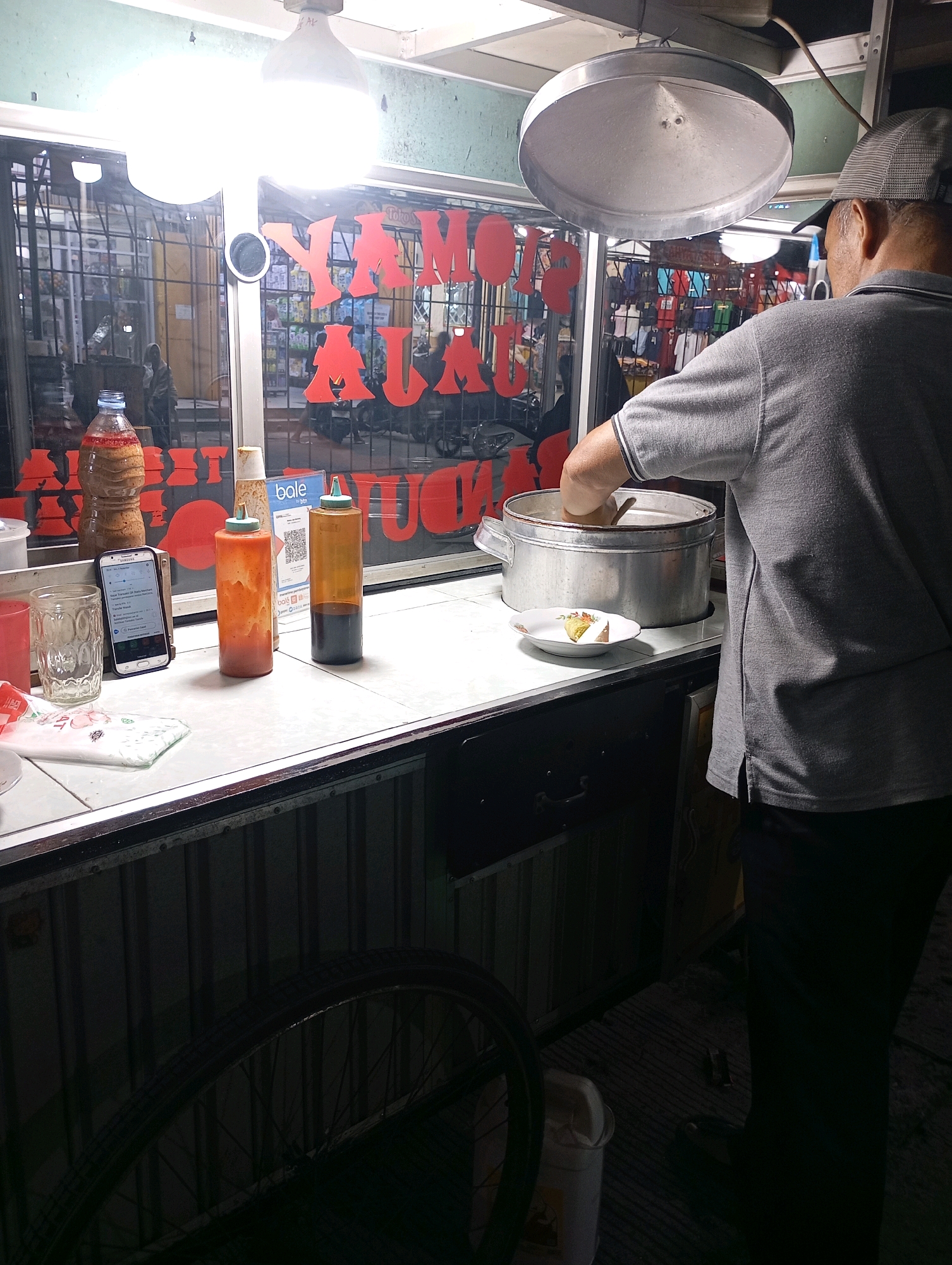 A man in a grey polo shirt and a baseball cap is standing behind a food stall in Babelan, Indonesia, and is in the process of serving food. The stall has a large metal pot, a small white plate with food on it, and two sauce bottles. A cell phone is propped up in front of the sauce bottles. Red lettering on the stall's window reads "TOMY AYAM". In the foreground, a bicycle wheel is visible. The overall lighting suggests it is nighttime.