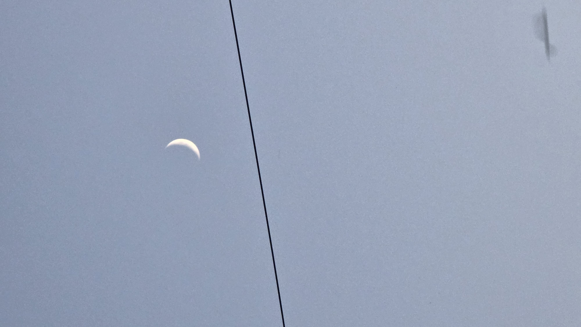 A thin crescent moon is visible in a pale blue sky. A dark horizontal line, likely a power line, cuts across the frame below the moon. The lower portion of the image contains a blurred, dark shape, possibly a cloud or an object in motion. The overall scene is minimalist and serene, with the moon as the focal point against the vast expanse of the sky.