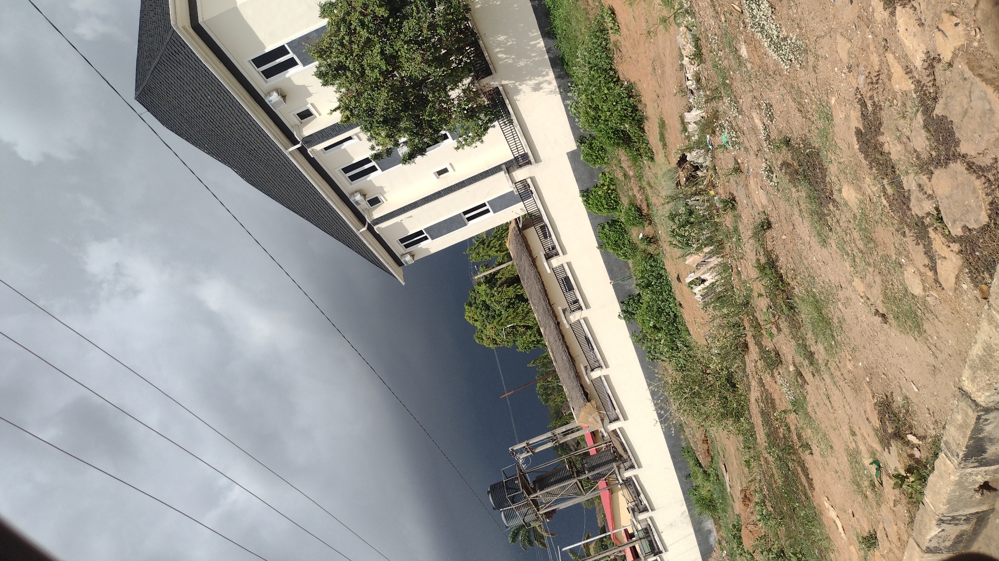 The image shows a multi-story building in Jos, Nigeria, under a cloudy sky. The building is cream-colored with dark-framed windows and a dark roof. A tree is visible near the building. Power lines are in the foreground. The ground is dirt and vegetation. A water tower or similar structure is visible closer to the bottom of the image, next to a smaller tree. The overall scene suggests an urban environment.
