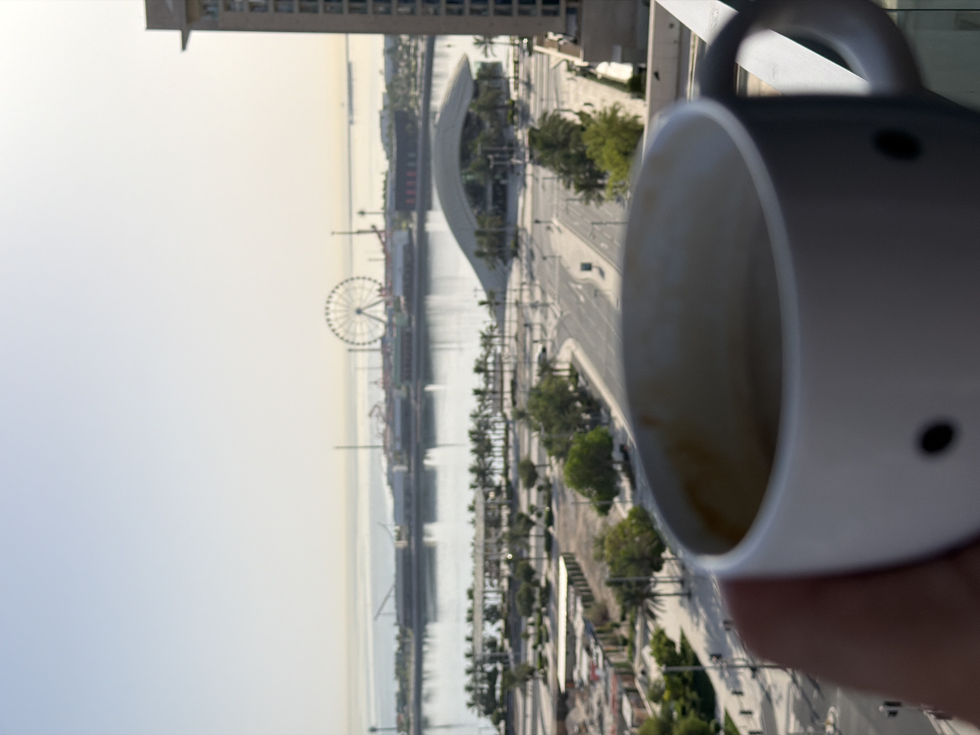 A person's hand holds a white coffee mug in the foreground, partially obscuring a blurry, sunlit cityscape in the background. The mug has two small black dots resembling eyes on its side. Beyond the mug, a body of water separates a developed waterfront from the open sea. On the waterfront, a Ferris wheel and several low-rise buildings are visible. The sky is a pale, hazy blue. The city appears to be a resort or entertainment district, likely located in a warm climate.