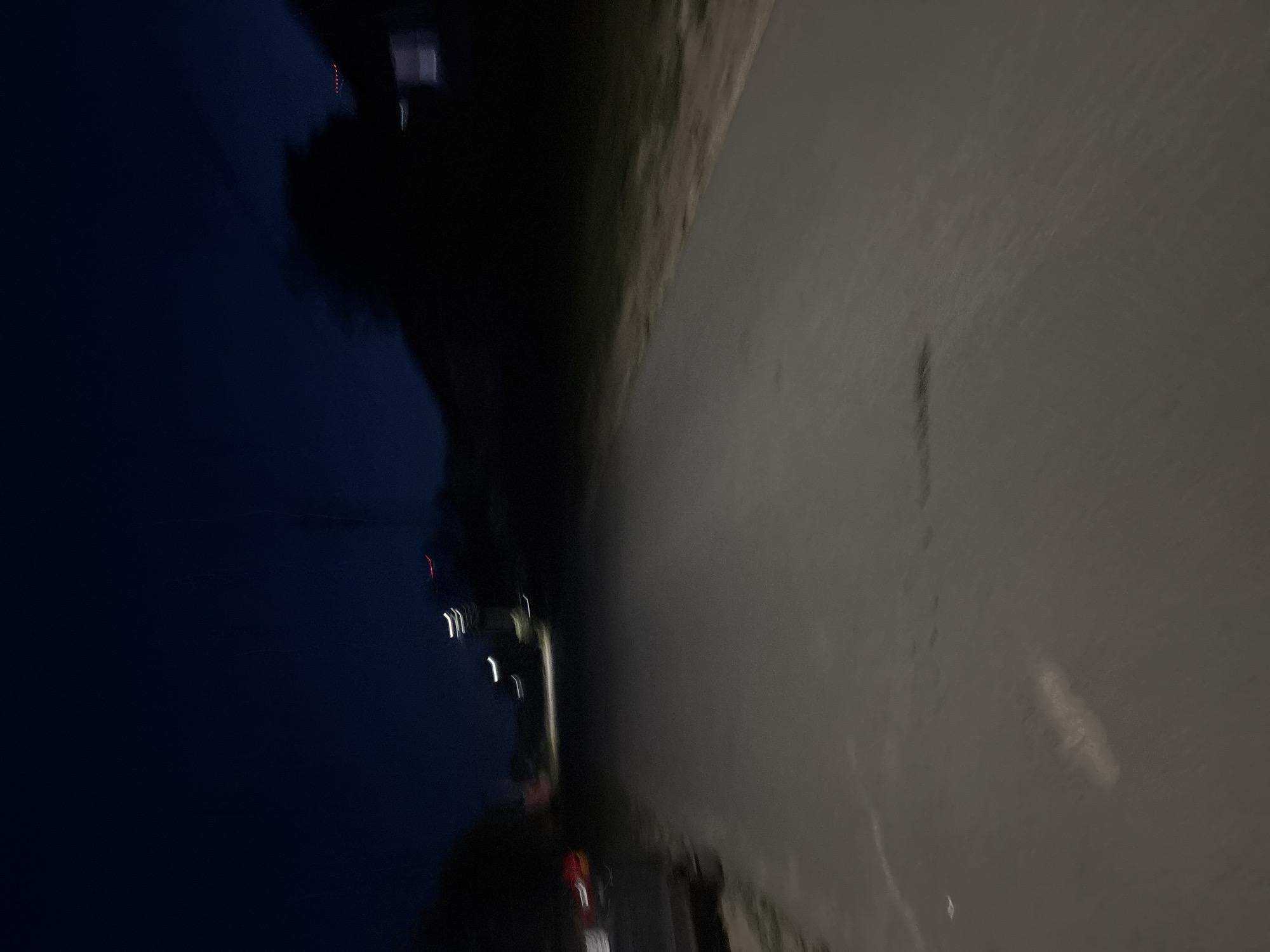 This nighttime image captures a paved road extending into the distance in the city of Unknown, Unknown. The scene exhibits significant motion blur, making precise identification challenging. The sky above is a dark blue-black. In the mid-distance, multiple light sources appear as blurred streaks: several are white or yellowish, and one is distinctly red, suggesting artificial illumination from streetlights or vehicles.
On the left side of the road, the rear portion of a dark-colored vehicle, possibly a car or SUV, is partially visible, displaying faint red taillights. Beyond the vehicle, a dark, indistinct mass, likely vegetation, is present. On the right side of the road, a strip of dark ground cover, consistent with grass or low vegetation, parallels the pavement. Further along the right, a dark silhouette of a structure, possibly a house, is discernible, featuring a single faint, rectangular light emanating from a window. The road surface itself is dark grey, showing a few indistinct lighter marks or areas of wear. No individuals or specific actions are discernible within the frame.