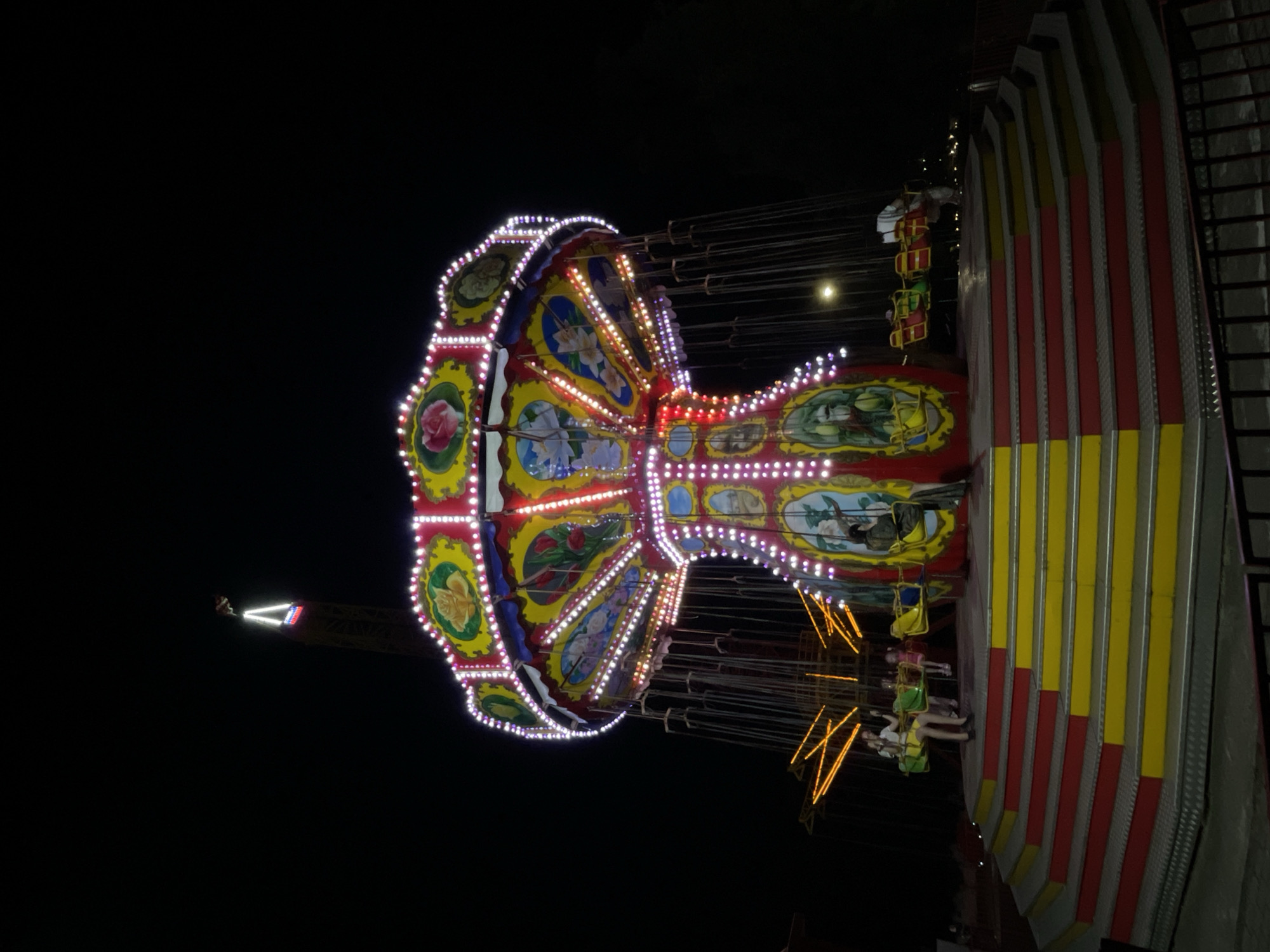 A brightly lit amusement ride, identified as a swing carousel, is the central focus of the image, photographed at night. The ride is adorned with numerous colorful lights, primarily white and pink, which illuminate its ornate structure. The carousel features painted panels depicting floral motifs and what appear to be figures. Several gondolas, painted in red and yellow, hang from the ride's arms, with a few people seated in them. A tiered platform with red and yellow steps leads up to the ride's boarding area. In the background, a tall structure with a triangular light at its apex is visible against the dark sky, suggesting a larger amusement park setting. The scene is devoid of any specific Izhevsk landmarks, and the context of the image being in Izhevsk, Russia, cannot be visually confirmed.