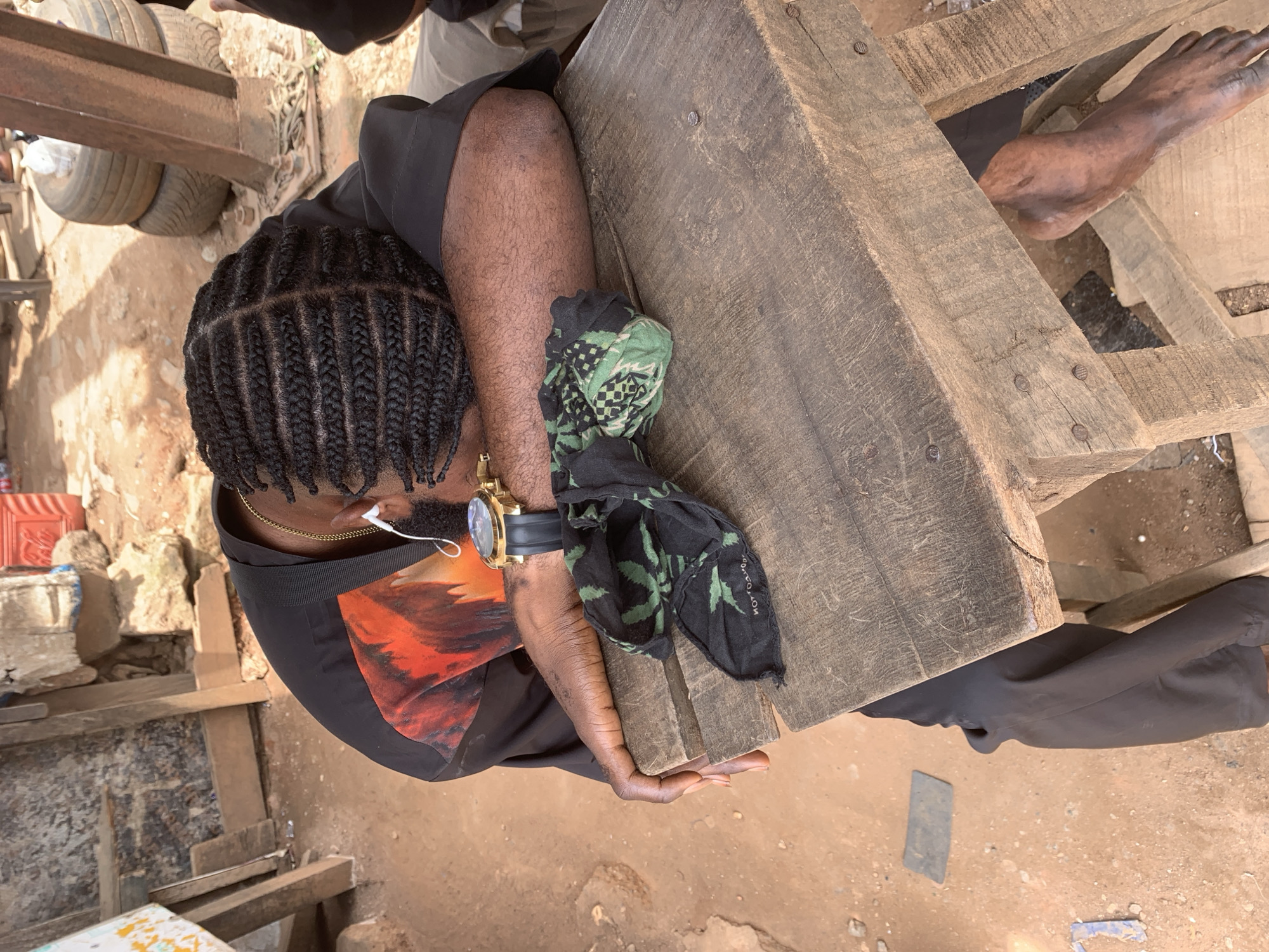 A person with braided hair, wearing a black shirt with an orange and black design, a gold watch, and white earbuds, is resting their head on their arms on a wooden table. A black cloth with a green cannabis leaf pattern is draped over the table. The scene appears to be outdoors, possibly in an informal market or workshop, given the dusty ground, stacked tires, and rustic wooden structures in the background. The overall impression is one of weariness or a moment of rest in a potentially busy environment.