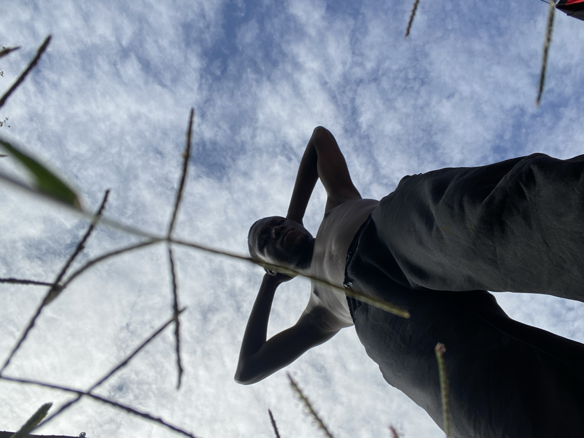 The image depicts a low-angle shot of a young Black person lying down outdoors, looking up at a cloudy sky. The subject is positioned centrally, with their head tilted back and hands behind their head, suggesting a relaxed or contemplative posture. They are wearing a light-colored tank top and dark pants. The foreground is partially obscured by blurred, thin blades of grass and plant stalks, creating a sense of depth and framing the subject. The sky is filled with white, wispy clouds against a bright blue background, indicating daytime. The perspective is from below, looking up towards the person and the sky, which emphasizes the vastness of the sky. No text is visible in the image. The setting appears to be a natural, outdoor environment, possibly a grassy area. The overall mood is serene and tranquil. The location is specified as Bukuru, Nigeria.