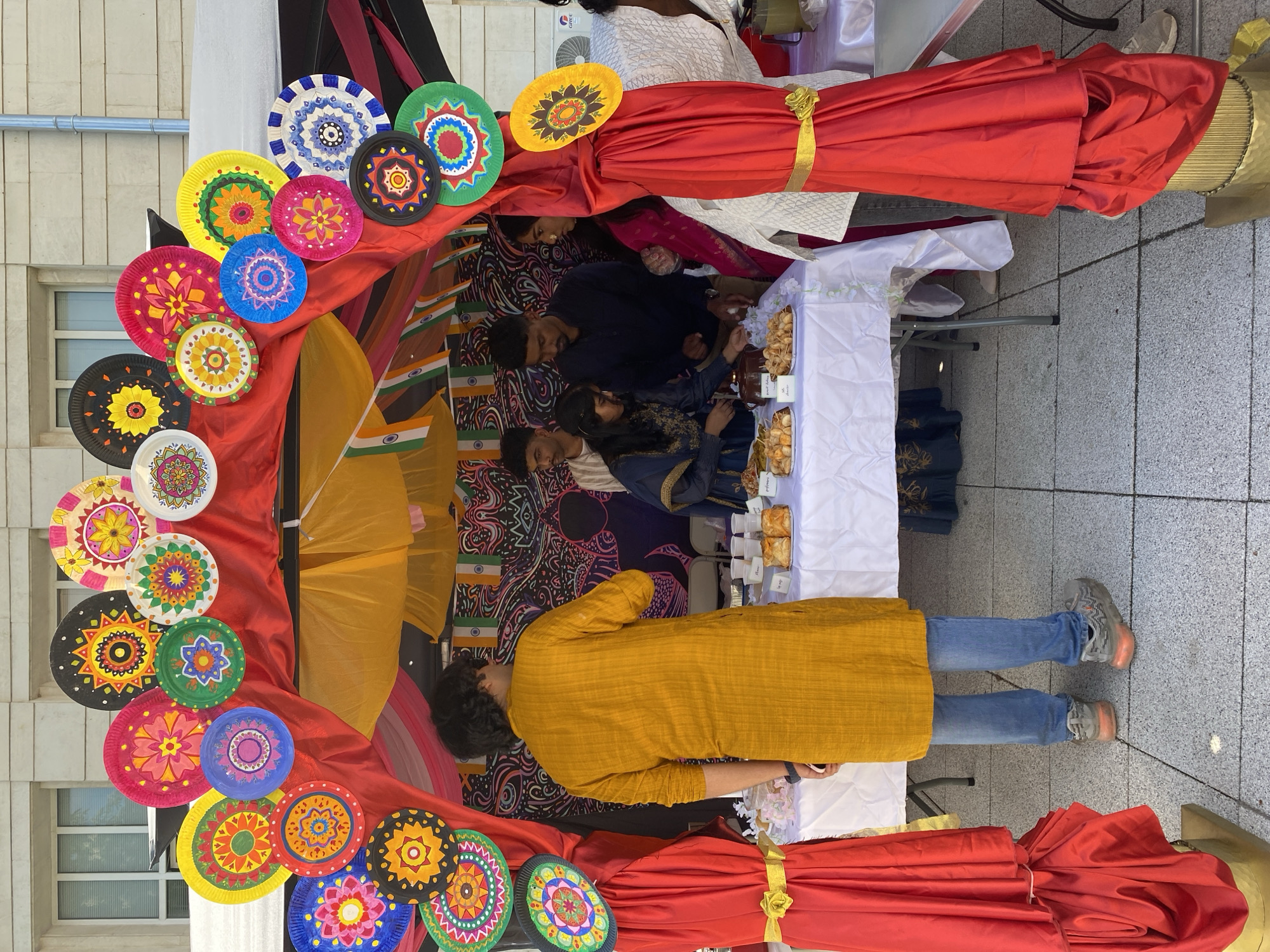A cultural booth adorned with colorful decorations is set up outdoors, possibly in the city of Pleven, Bulgaria. People are gathered around the booth, some standing and others seated behind a table. The table is covered with a white cloth and displays what appear to be food items or pastries. A person in a yellow top and jeans stands in front of the table, seemingly engaging with the items or people at the booth. The booth is decorated with a red drape, colorful paper plates arranged in a decorative pattern, and flags. The environment appears to be an open space near a building.