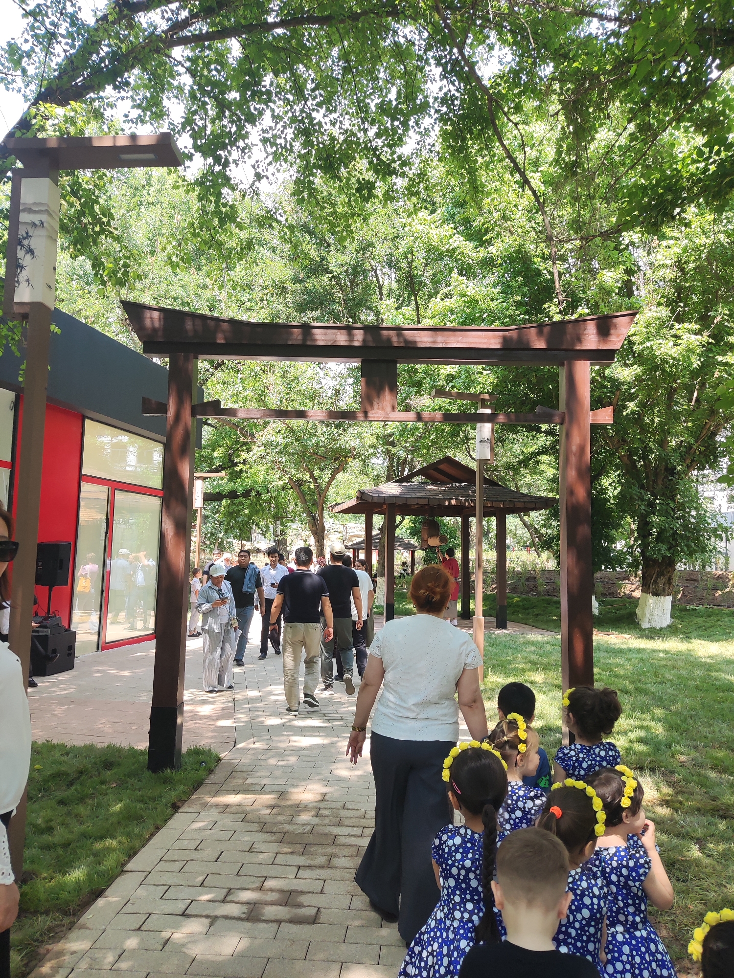In Shymkent, Kazakhstan, this image captures an outdoor scene featuring a pathway lined with trees and a Japanese-style torii gate. A group of people, including adults and several young girls in matching blue and white polka dot dresses and yellow flower crowns, are walking along the pathway. To the left, a modern building with red accents and large windows is visible, with a speaker system set up outside. The atmosphere appears to be festive, possibly indicating an event or gathering. The ground is paved with light-colored bricks, and lush green grass borders the path. The trees provide ample shade, suggesting a warm day.