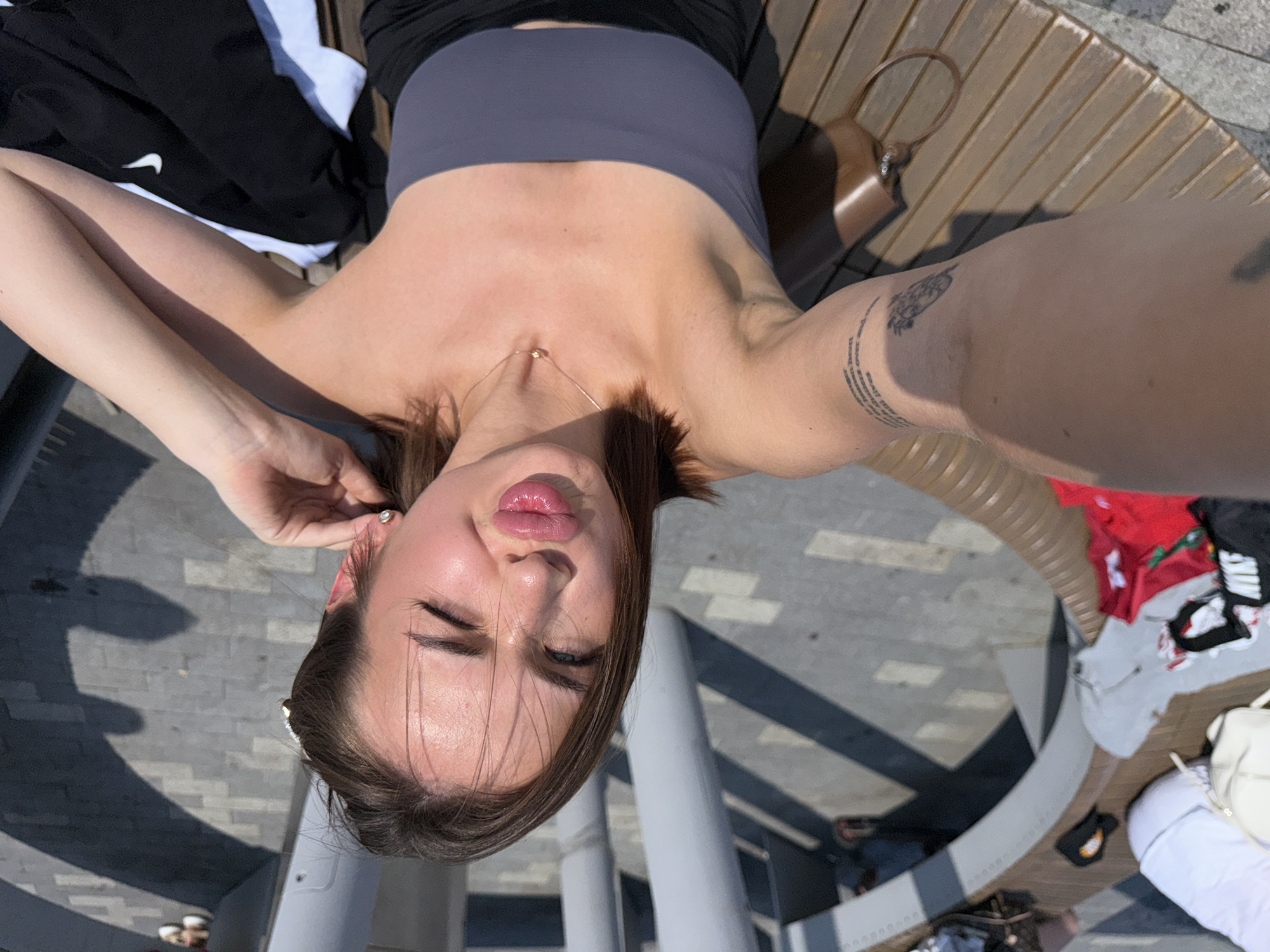 A woman with brown hair and fair skin is taking a selfie outdoors. She is wearing a grey strapless top and black shorts. She has a tattoo on her left arm and is wearing a necklace. She is sitting on a wooden bench. In the background, there are some large grey pillars and patterned paving stones. There are also some bags and clothing items on the bench. The location is the embankment of the Kama River in Perm, Russia.