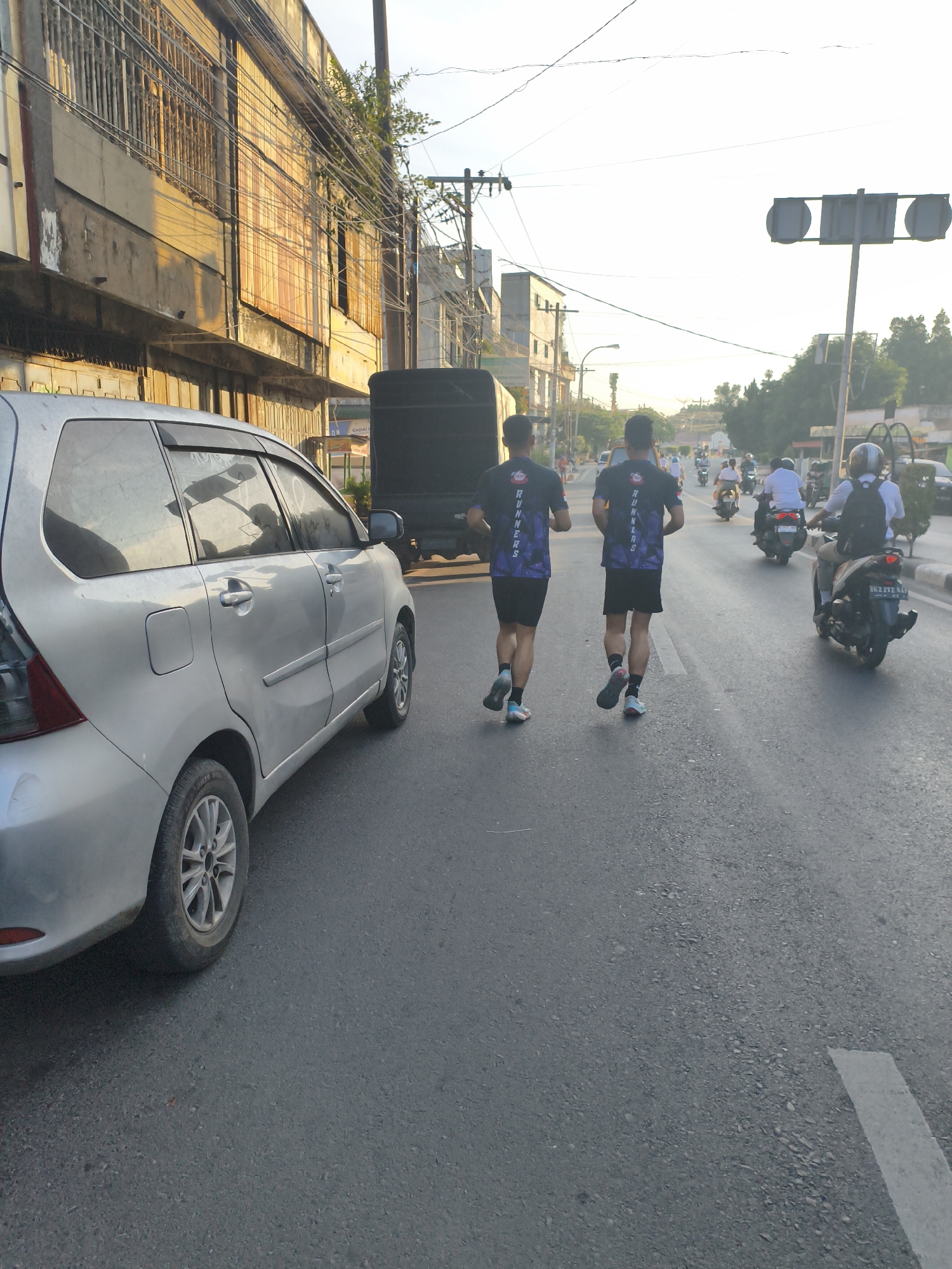 In the city of Tebing Tinggi, Indonesia, two individuals, likely participants in a running event, are depicted from behind as they jog along a paved road. They are both wearing black t-shirts with purple text that reads "RUNNERS" and black shorts. The road is somewhat busy, with several motorcycles in the background. One motorcycle is to the right of the runners, with a rider wearing a helmet and a white shirt, and another person on the back. Further down the road, other motorcycles and some vehicles are visible. To the left of the road, there is a silver minivan. In the background, there are buildings with weathered facades and exposed electrical wires running overhead. The sky is bright and appears to be late afternoon or early morning, casting a warm light on the scene. A traffic light is visible in the distance, mounted on a pole. The overall environment suggests an urban setting during a daytime activity.