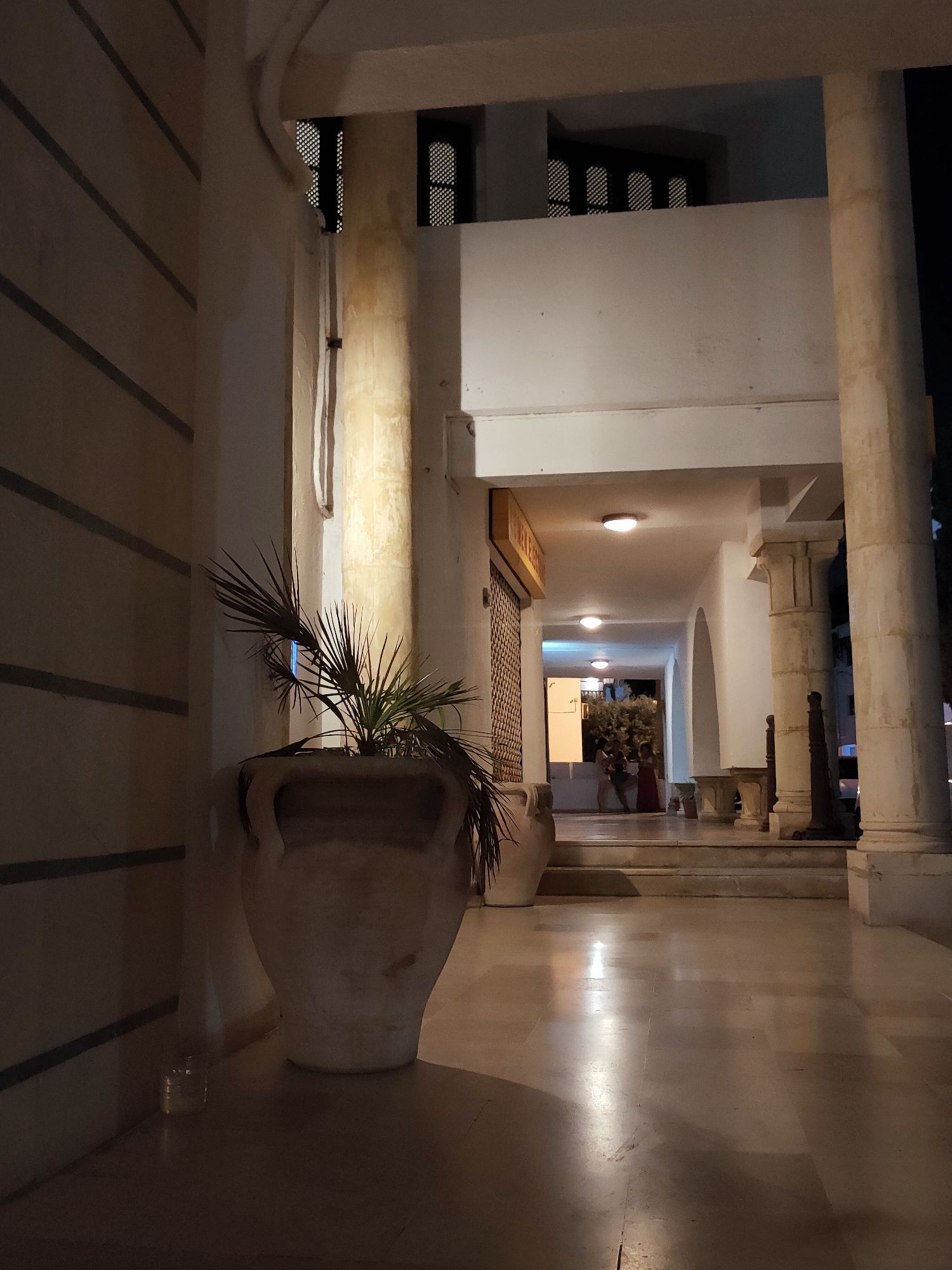 An exterior shot of a building's covered walkway at night in Tunis, Tunisia, shows large stone pillars and a tiled floor reflecting light from overhead fixtures. A large, light-colored ceramic pot with a plant stands on the left side of the frame. In the distance, a group of people are visible near an entrance, illuminated by interior lights. The architecture suggests a historical or traditional style.